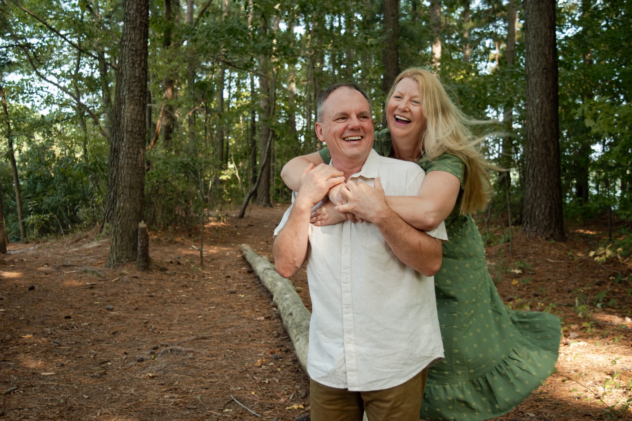A man and woman laughing outdoors among trees, with the woman hugging the man from behind.