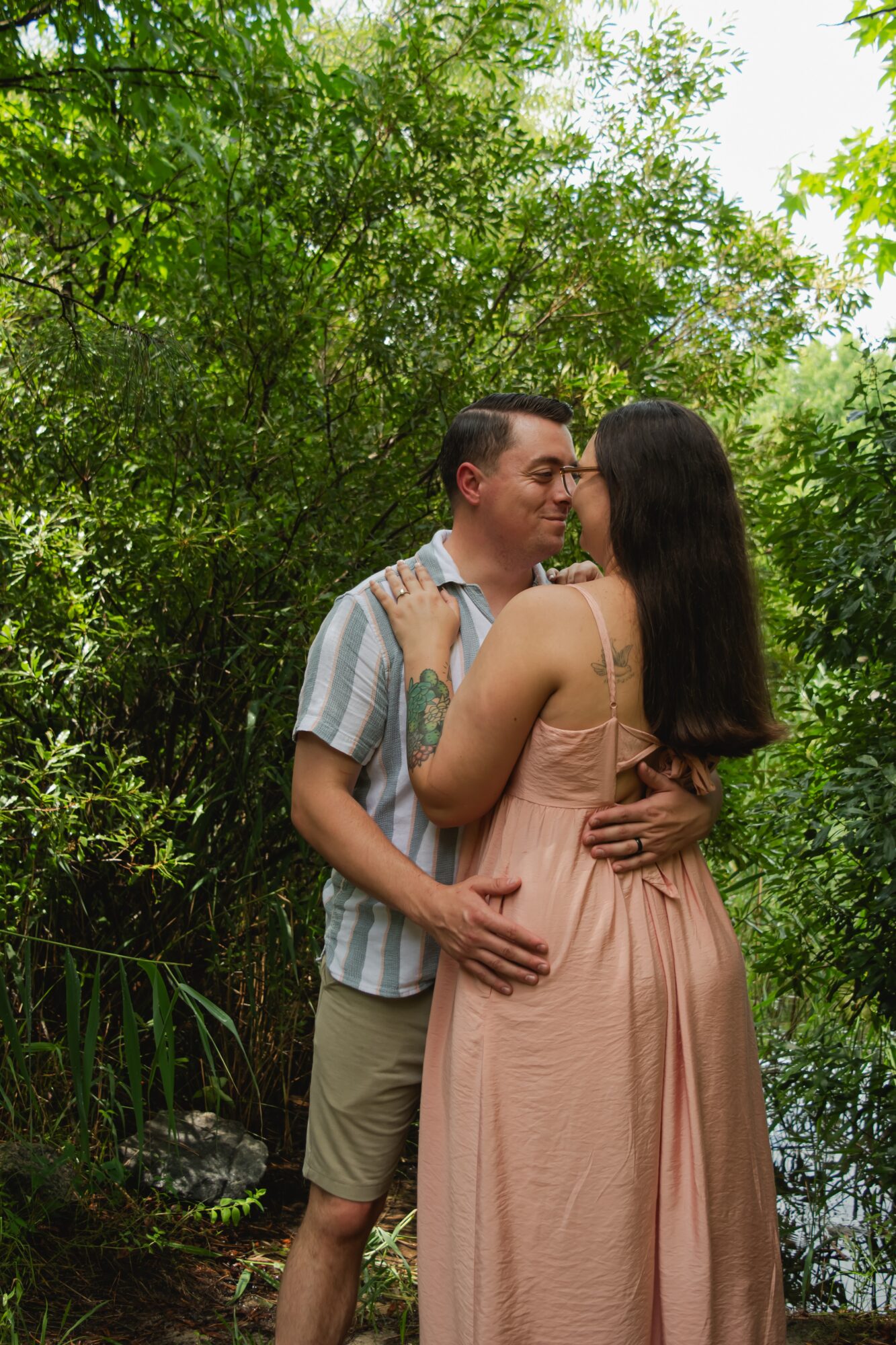 A couple stands close together outdoors surrounded by green trees, about to kiss, with the woman in a pink dress and the man in a striped shirt.