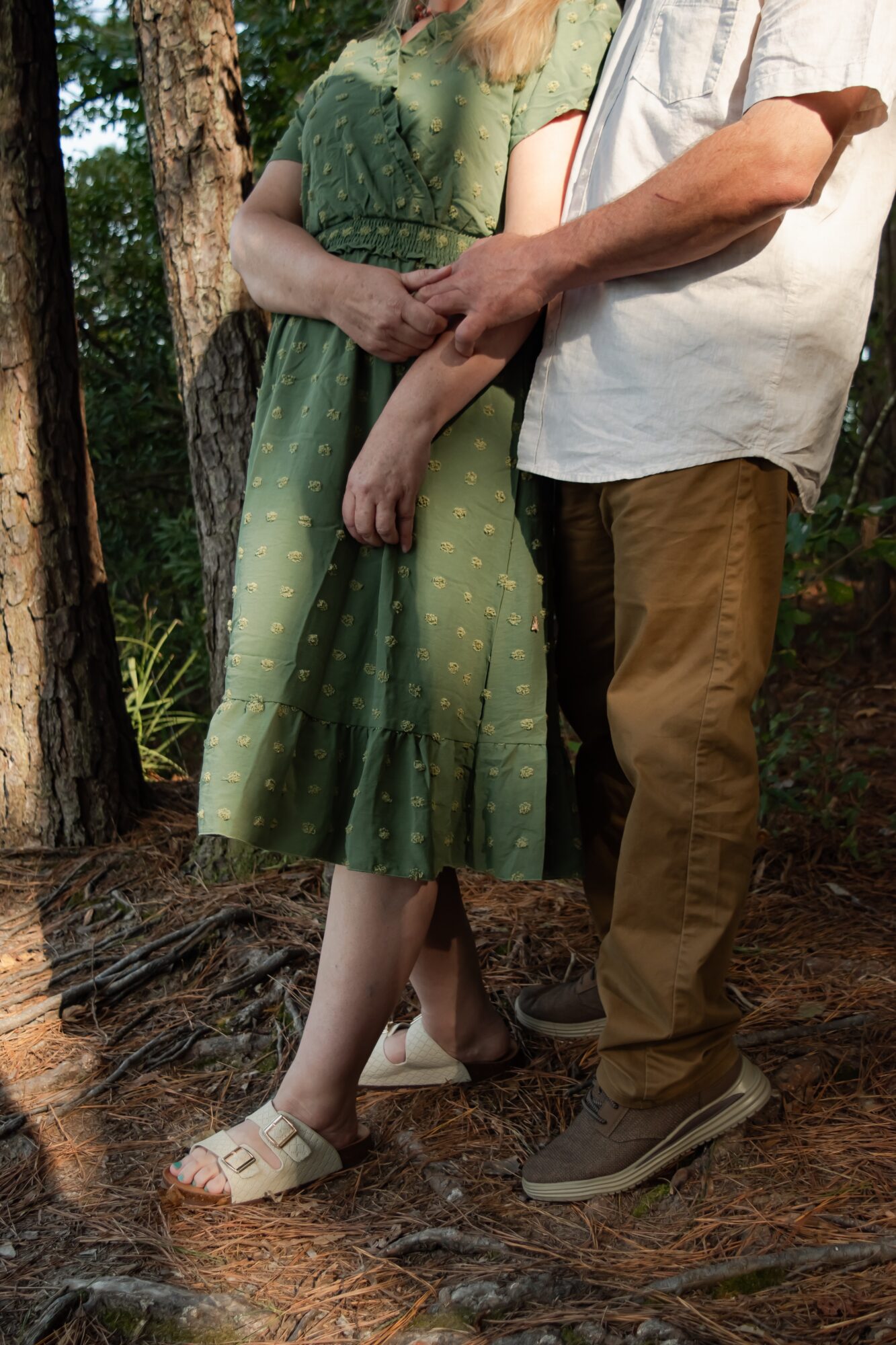 Two people standing outdoors, one with a green dress and the other with brown pants, holding hands.
