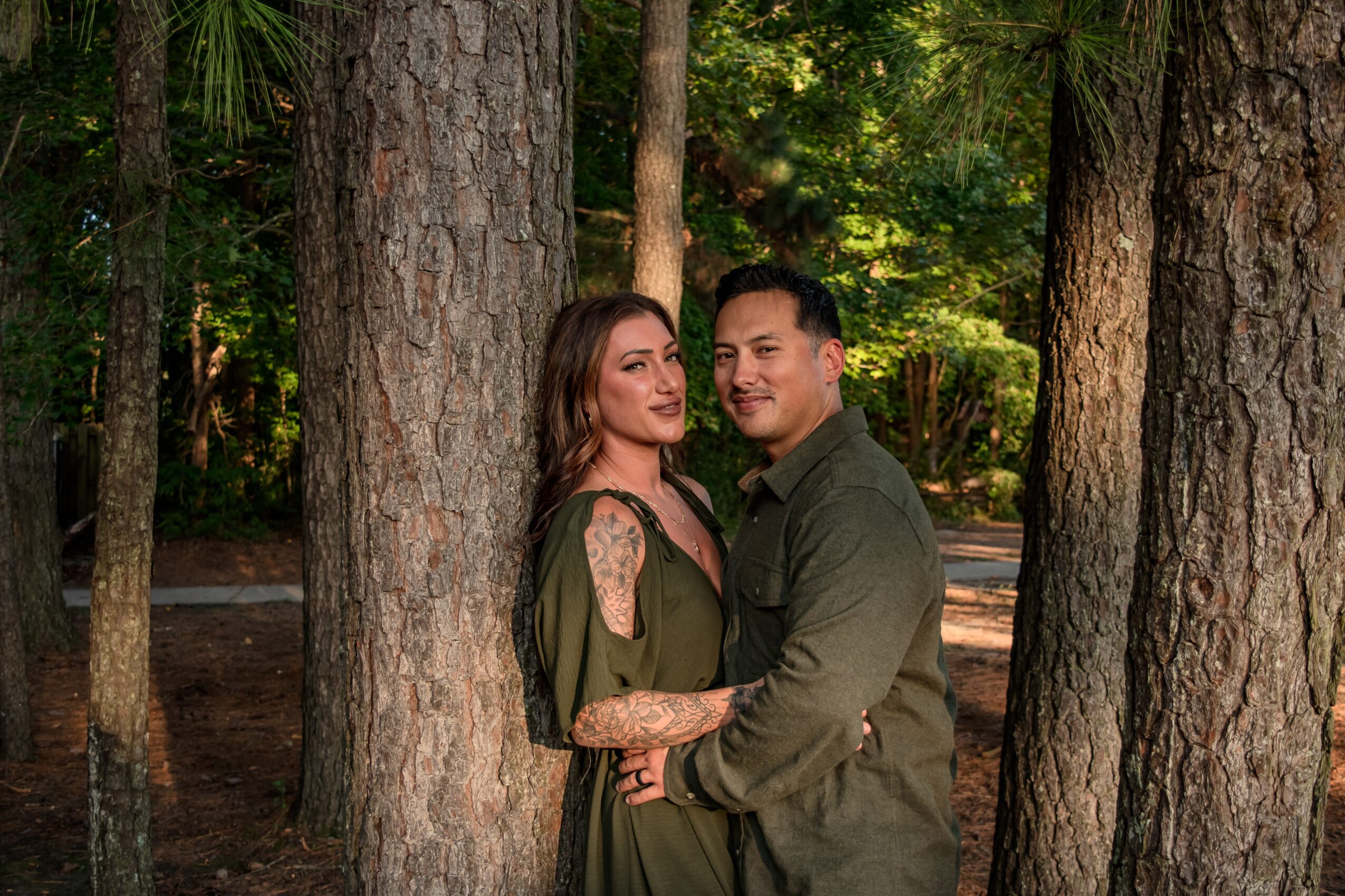 A couple stands between trees in a forest, smiling and embracing each other, surrounded by green foliage.