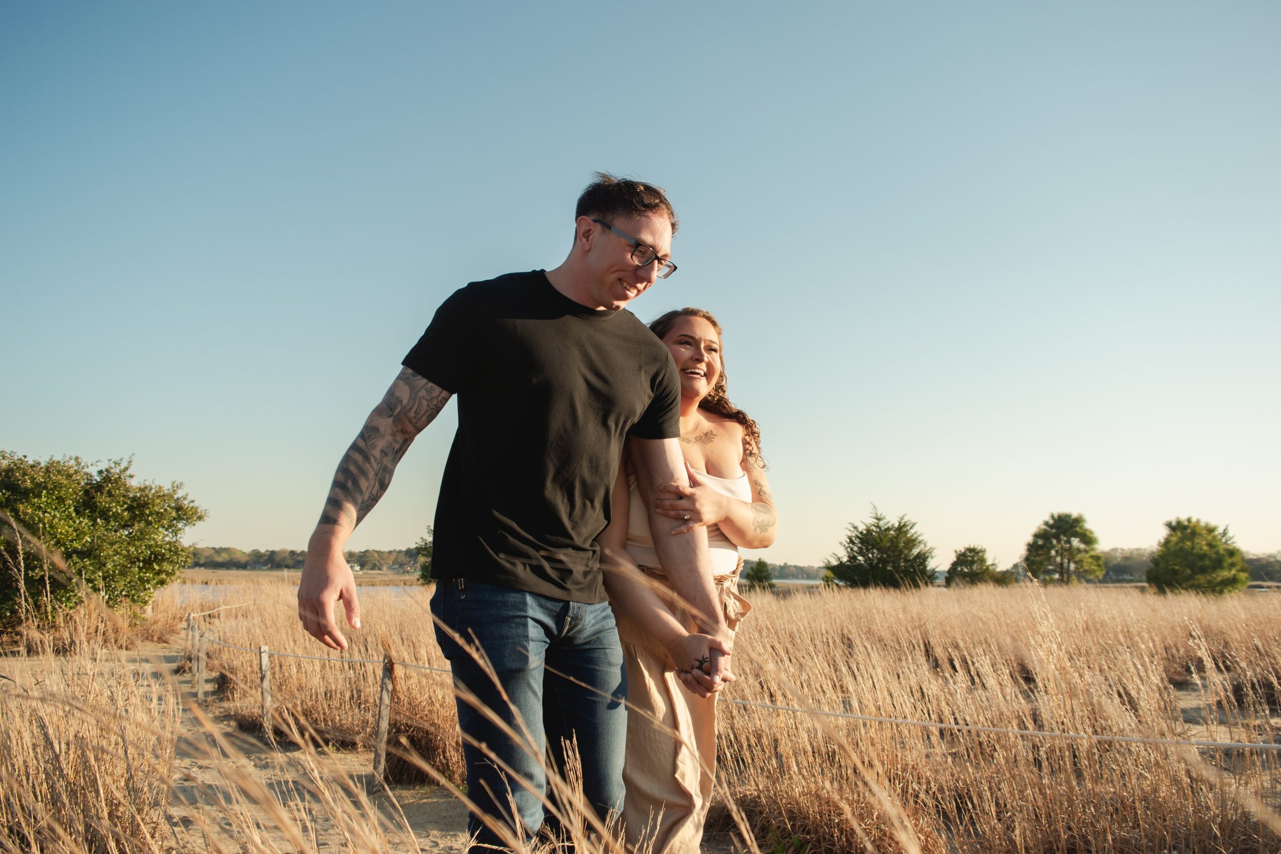 A man and woman walk through a field holding hands, with a clear sky and trees in the background.