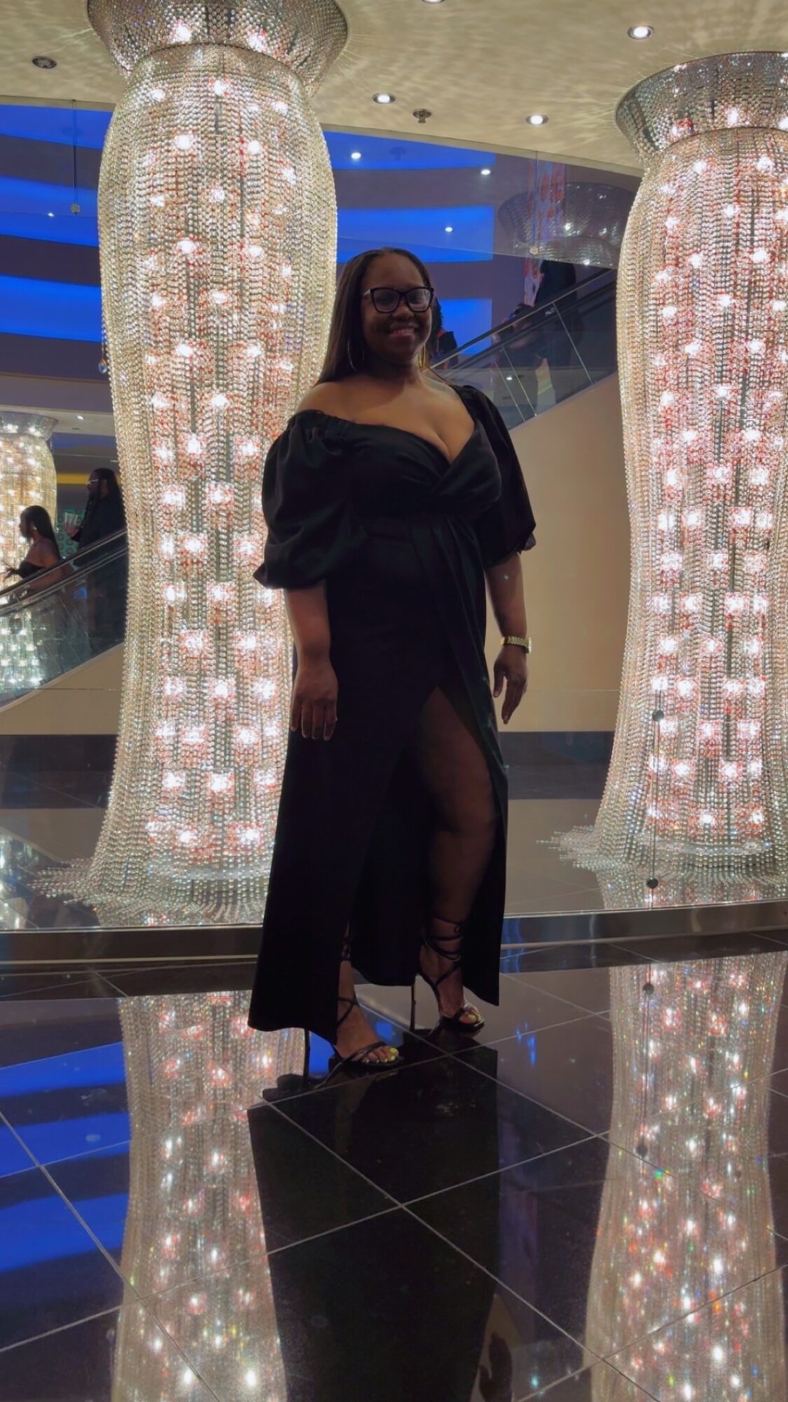 Woman in black dress standing between two illuminated, decorative columns with reflections on the shiny floor.