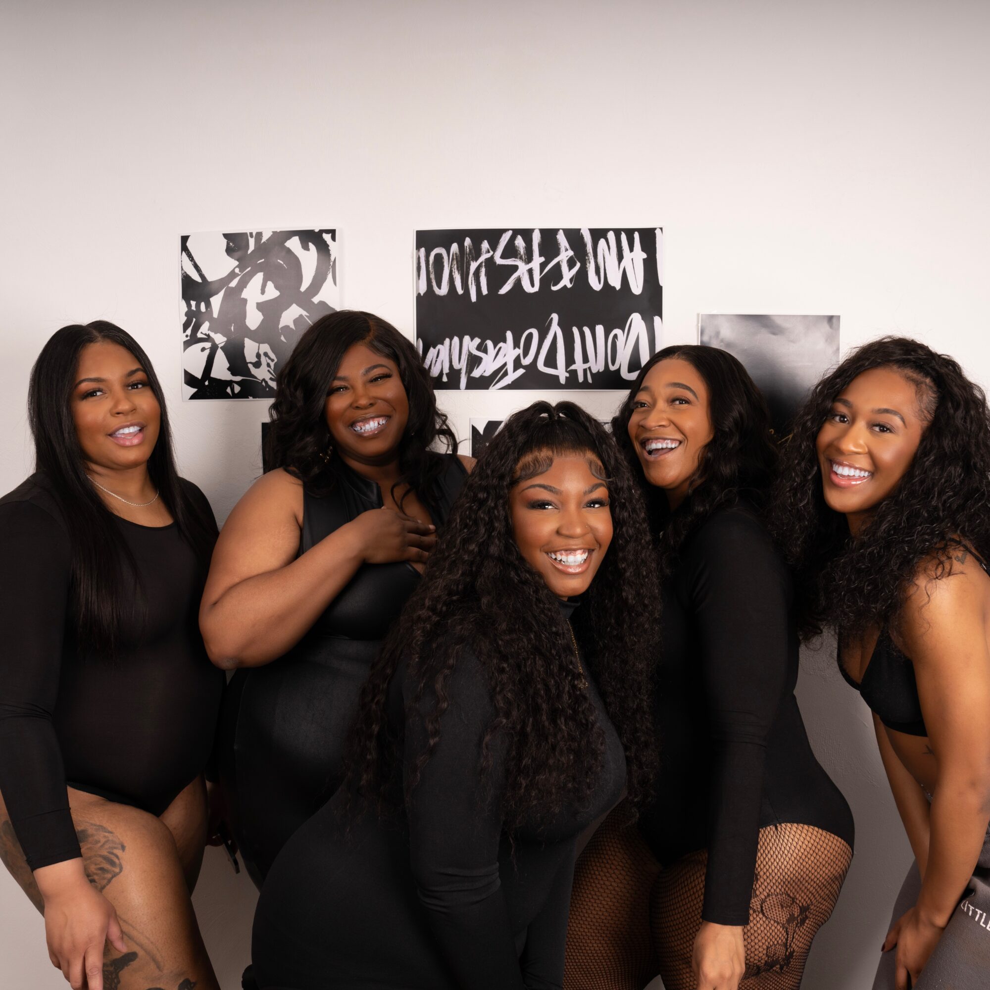 Five women smiling and posing together indoors, with abstract black and white artwork on the wall behind them.