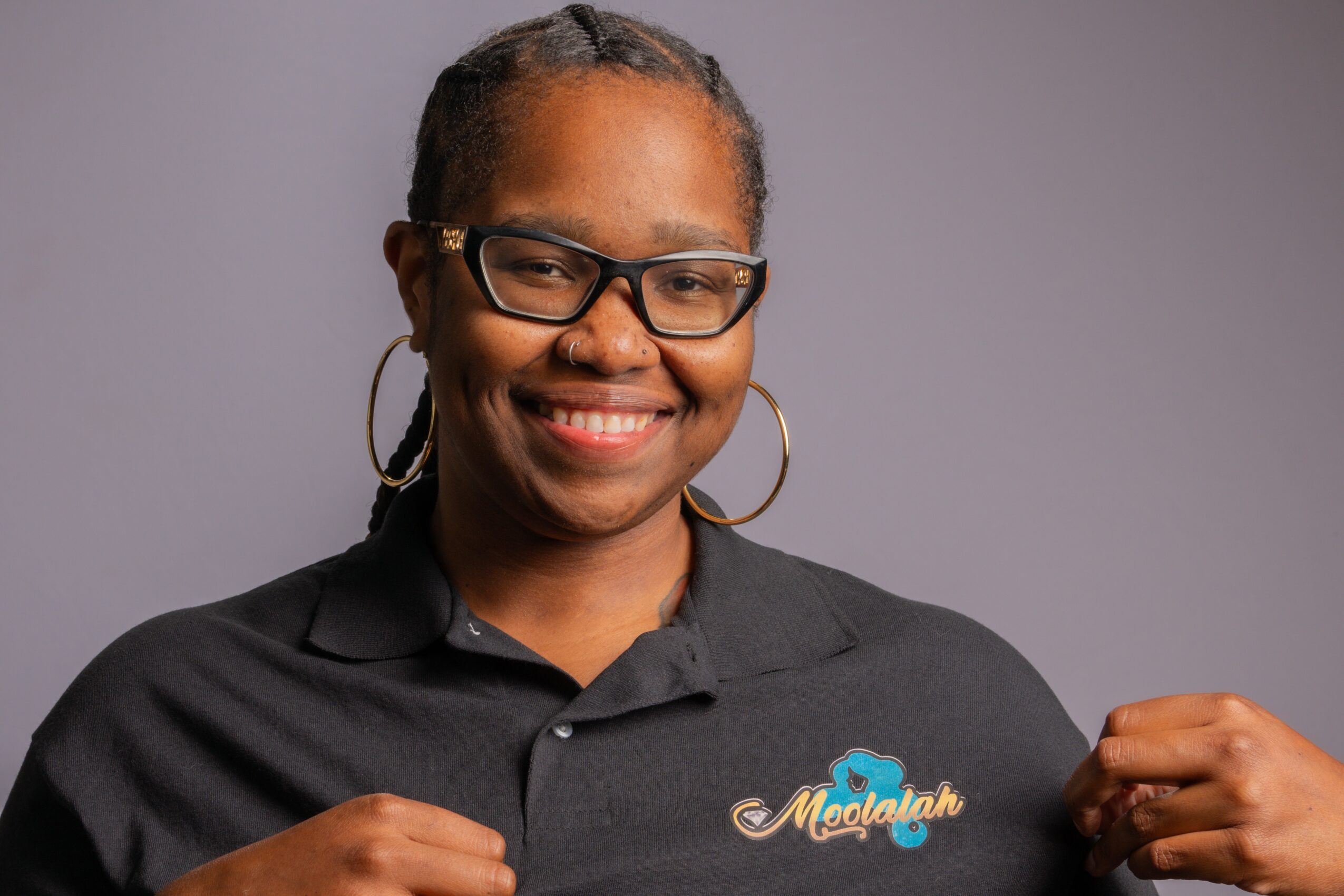 Smiling woman with glasses and hoop earrings points to a sign that says 'Mogul' against a plain background.