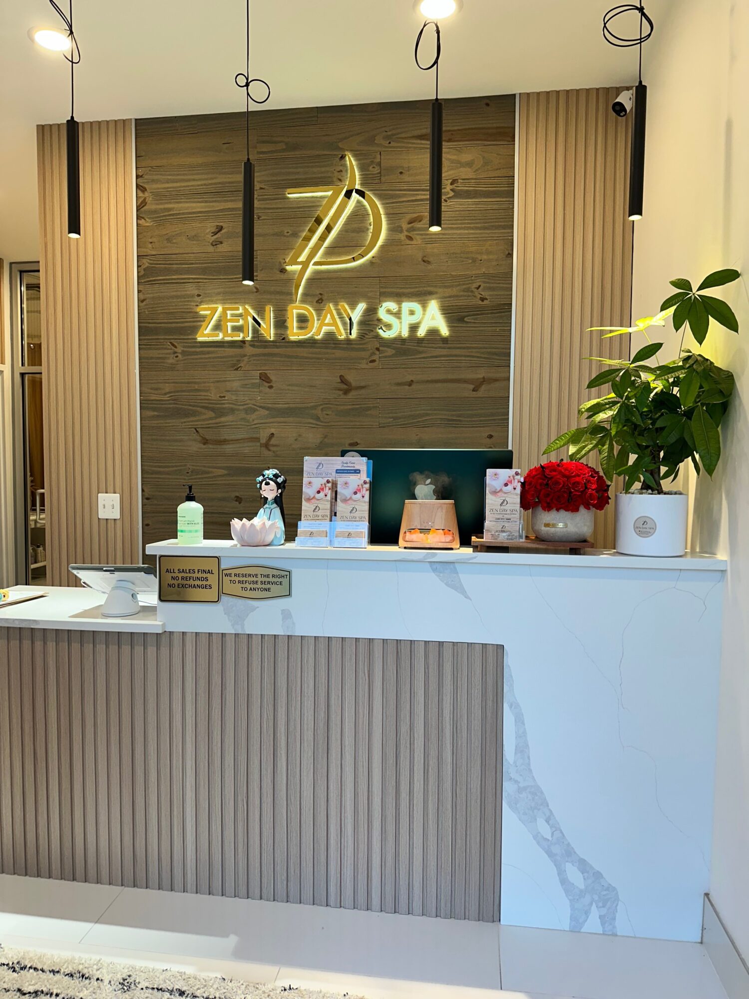 Reception desk with plants, spa products, and a sign reading 'ZEN DAY SPA' on a wooden wall, viewed from the side.
