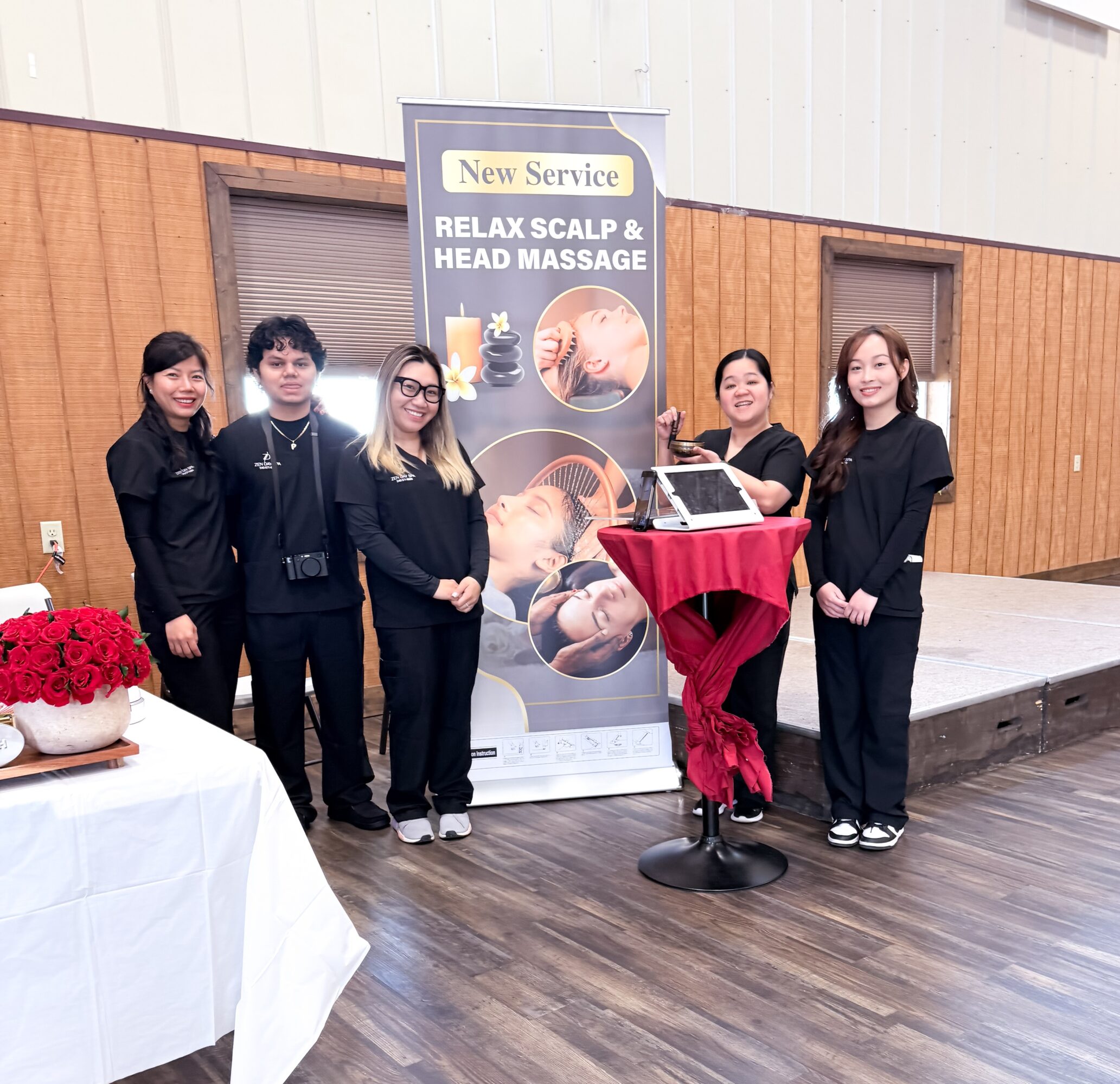 Five people stand next to a promotional banner for scalp and head massage services, with a table of red flowers nearby.