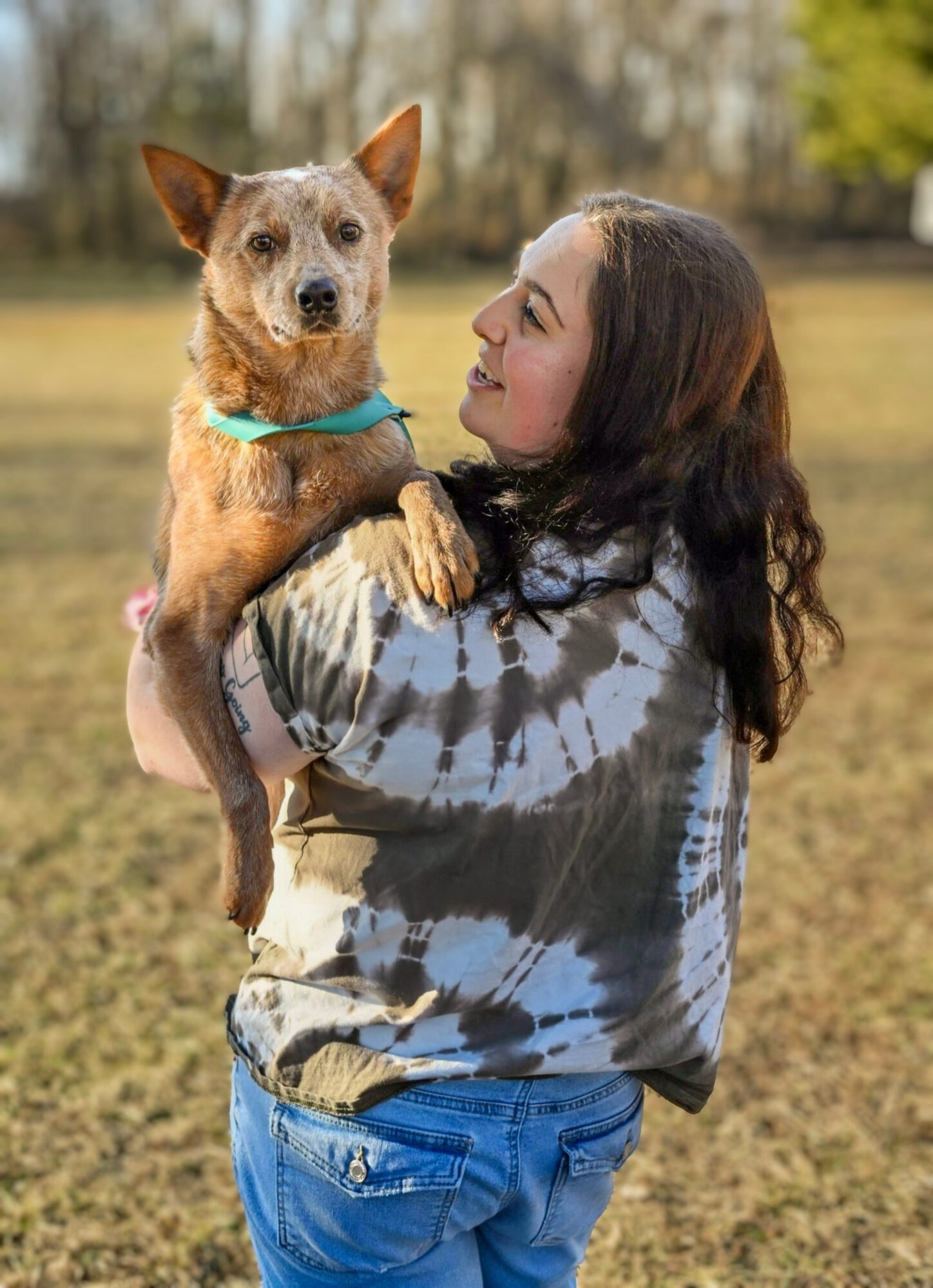 Woman holding a small dog outdoors in a grassy area, trees in the background, woman smiling at the dog.