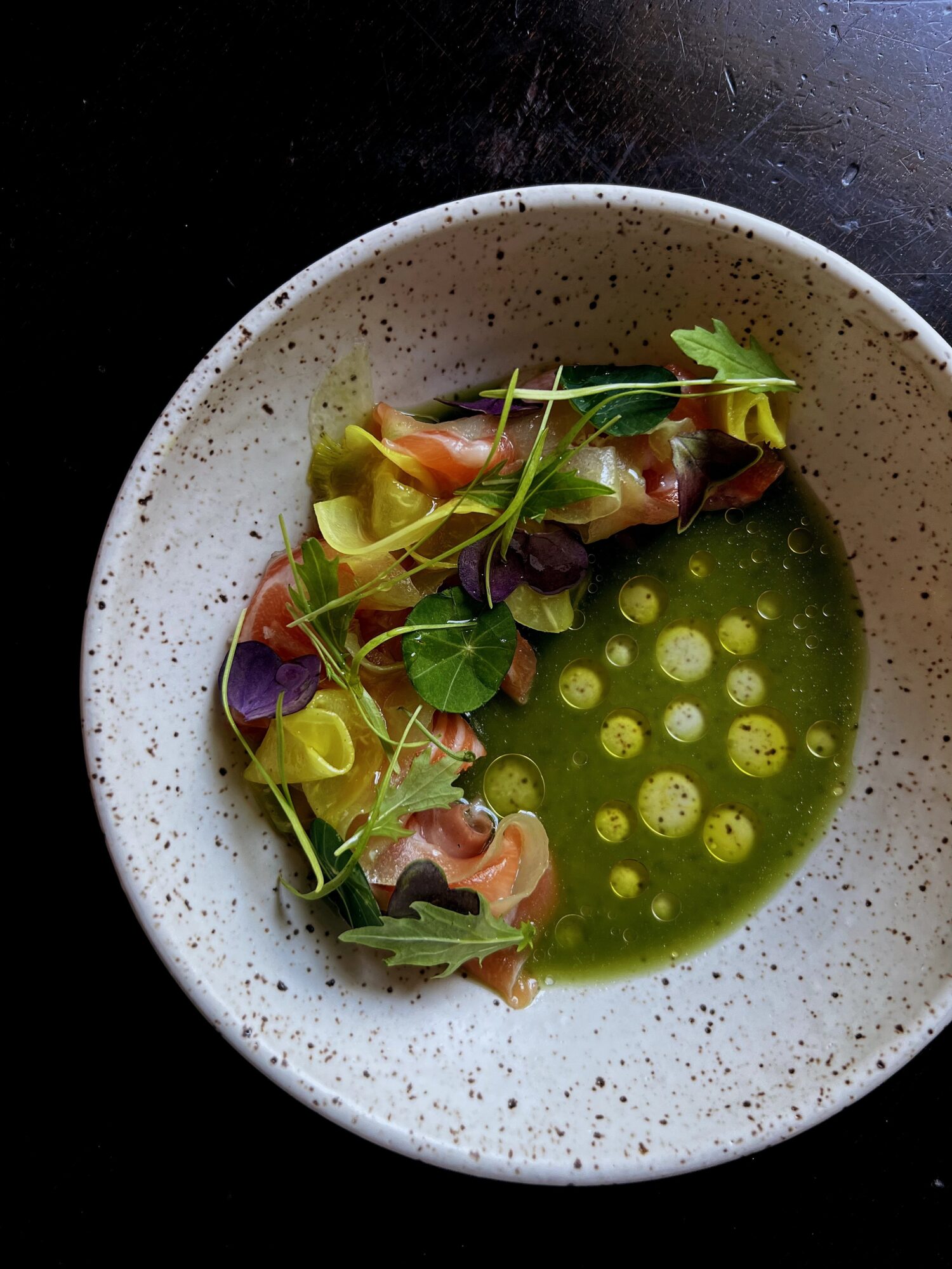 Dish with rolled food, green sauce, and microgreens in a speckled bowl on a dark surface.