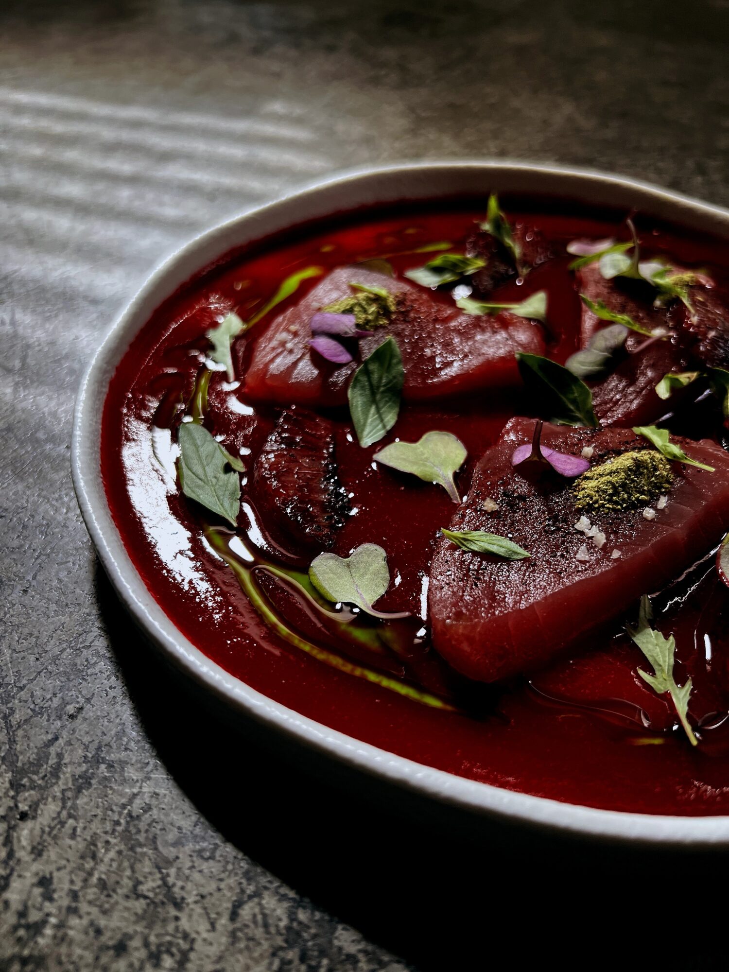 Bowl of cooked meat in red sauce garnished with herbs and greens, on a dark textured surface.