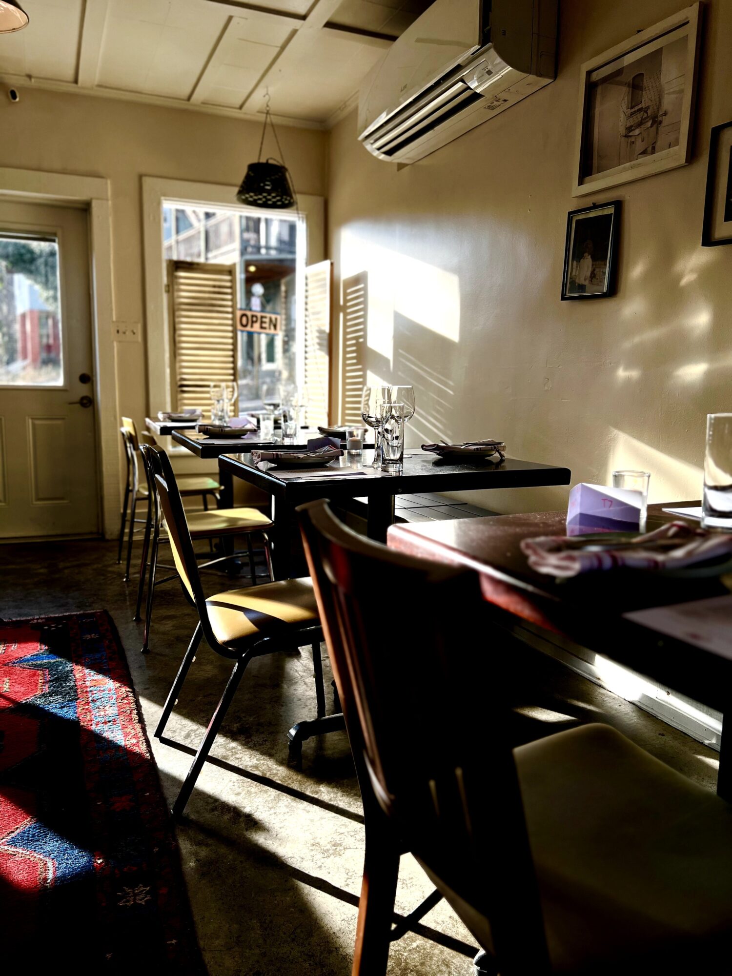 Dining area with table set for meals, sunlight streaming through window, framed pictures on wall, and chairs around the table.