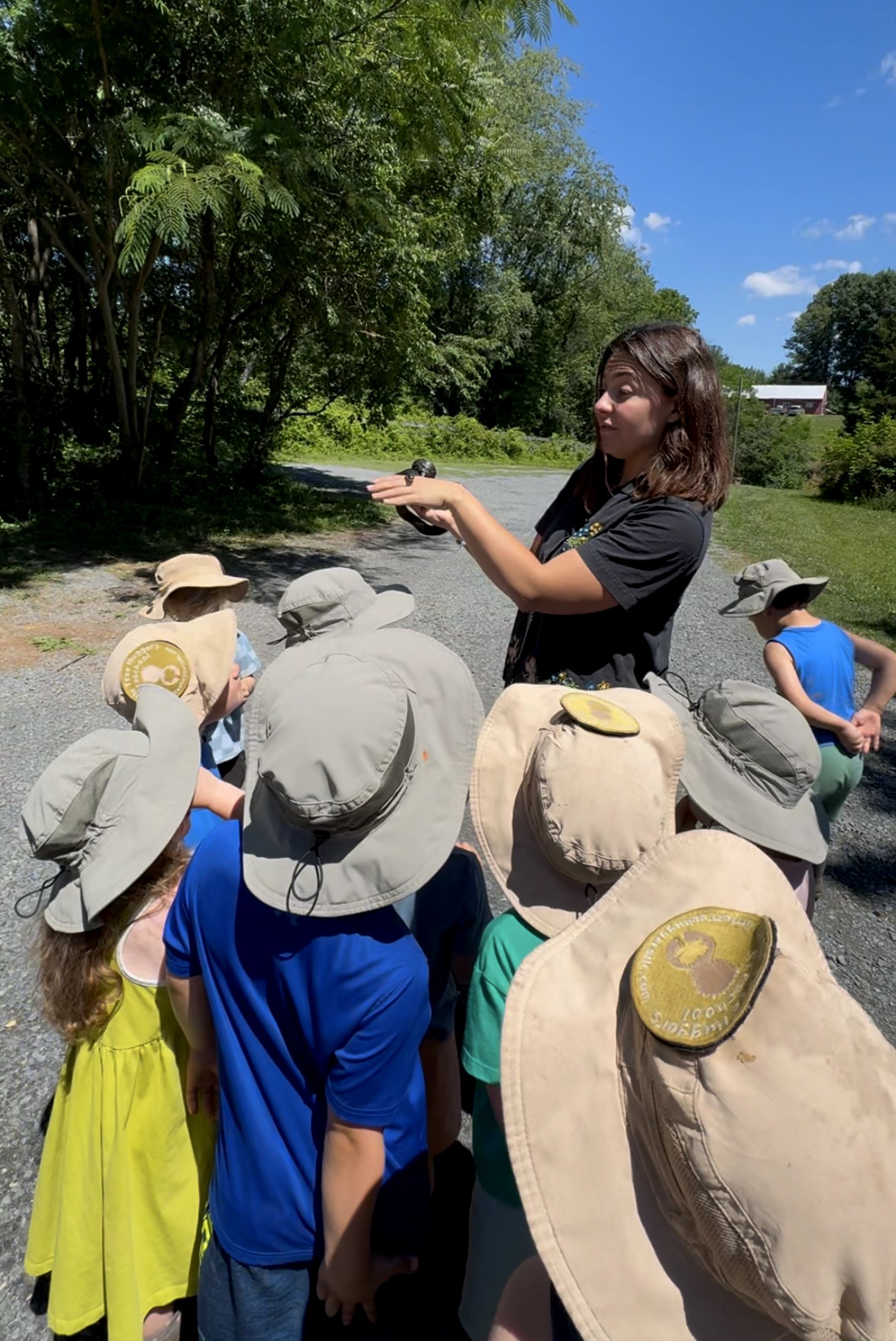 Woman demonstrating something to children outdoors, children wearing hats, trees and blue sky in background.