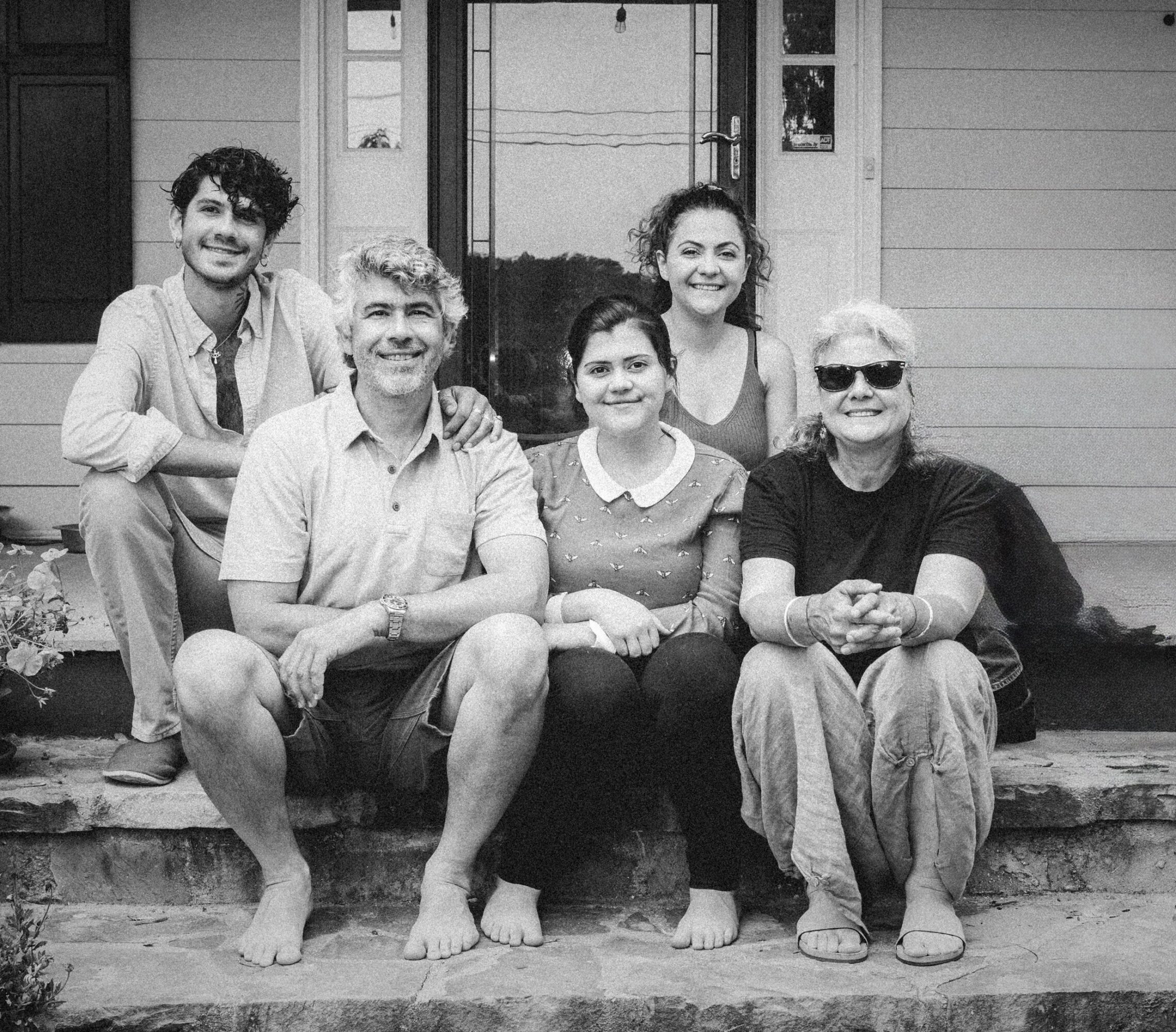 Group of six people sitting and standing on porch steps in front of a house, smiling, black and white photo.