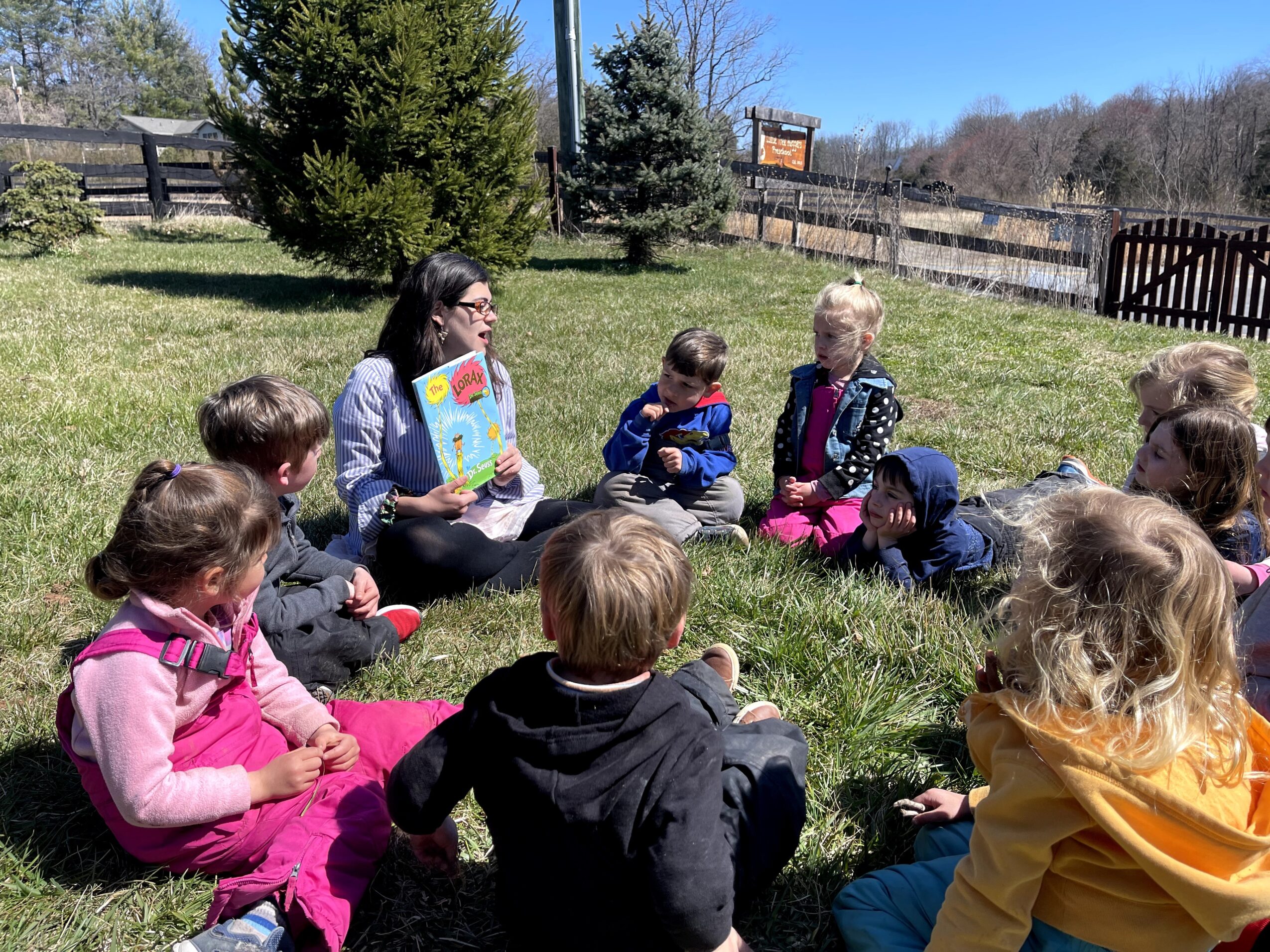Group of children and an adult sitting on grass outdoors, engaging in a discussion or activity, with trees and a fence in background.