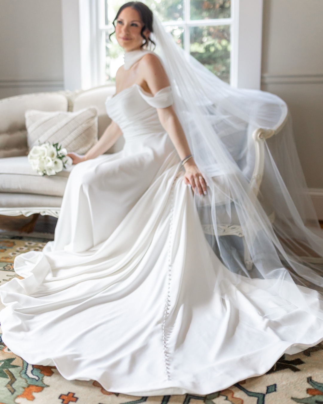 Bride in a white wedding dress sitting on a sofa, holding a bouquet, with a veil flowing behind her.