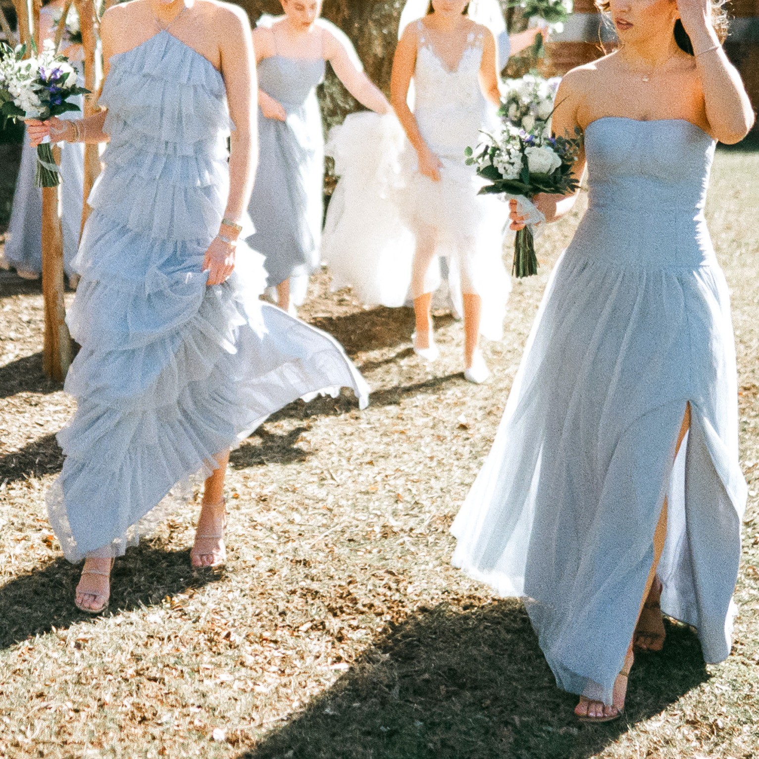 Group of women in light blue dresses walking outdoors, holding flowers, with sunlight and grass visible.