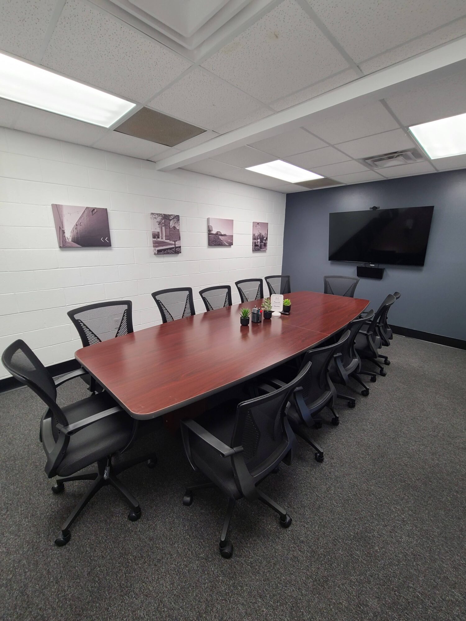 Conference room with a long table, chairs, a wall-mounted TV, and framed pictures on the wall.