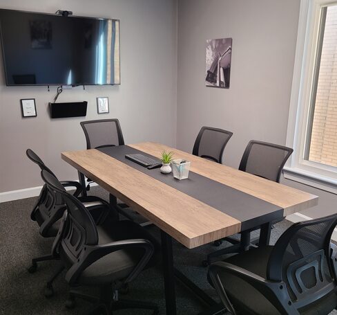 Conference room with a wooden table, six chairs, a wall-mounted TV, and a window, with a plant and notebooks on the table.
