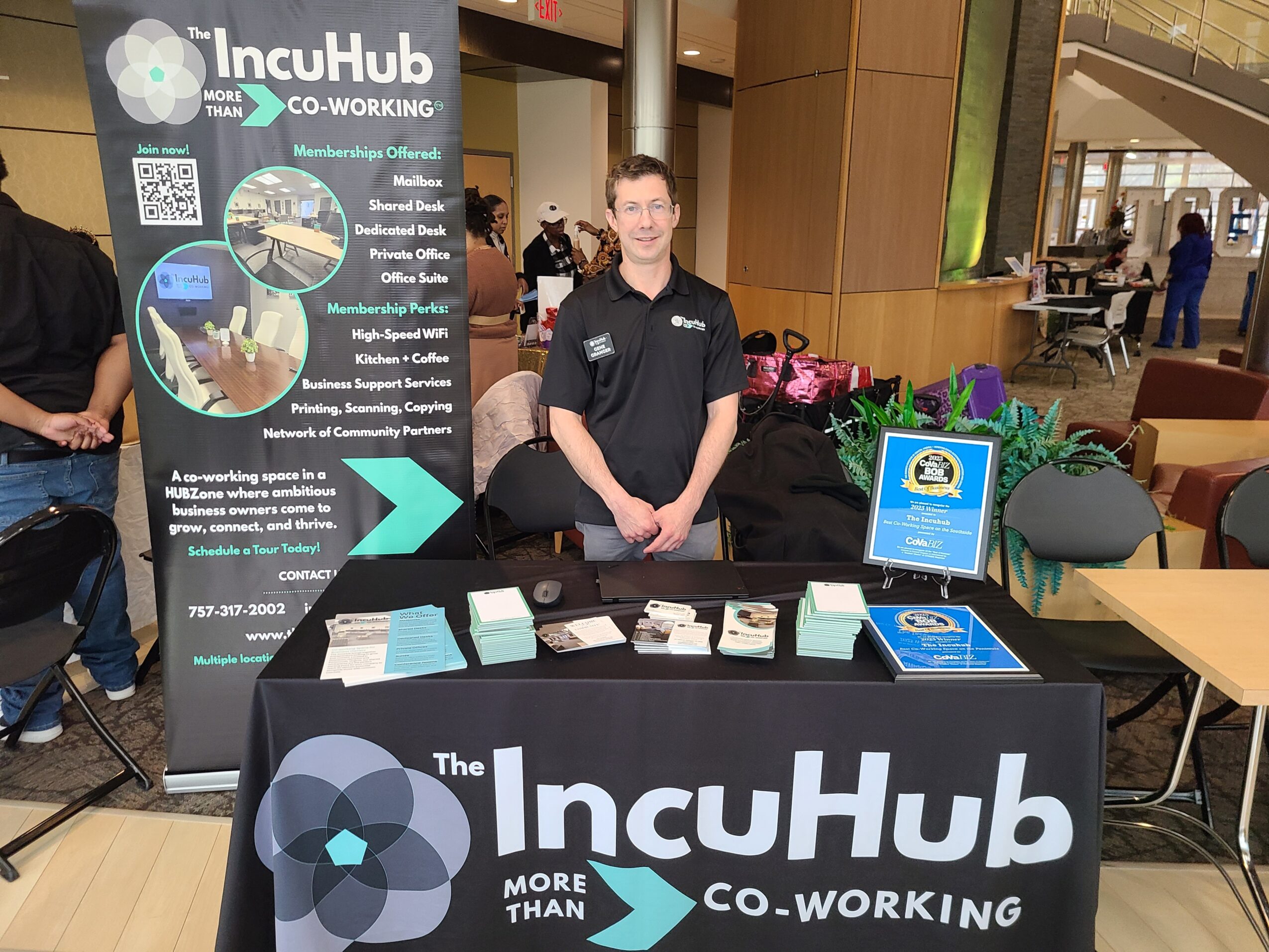 Man standing behind a table with promotional materials at an event, with a banner and a sign on the table.