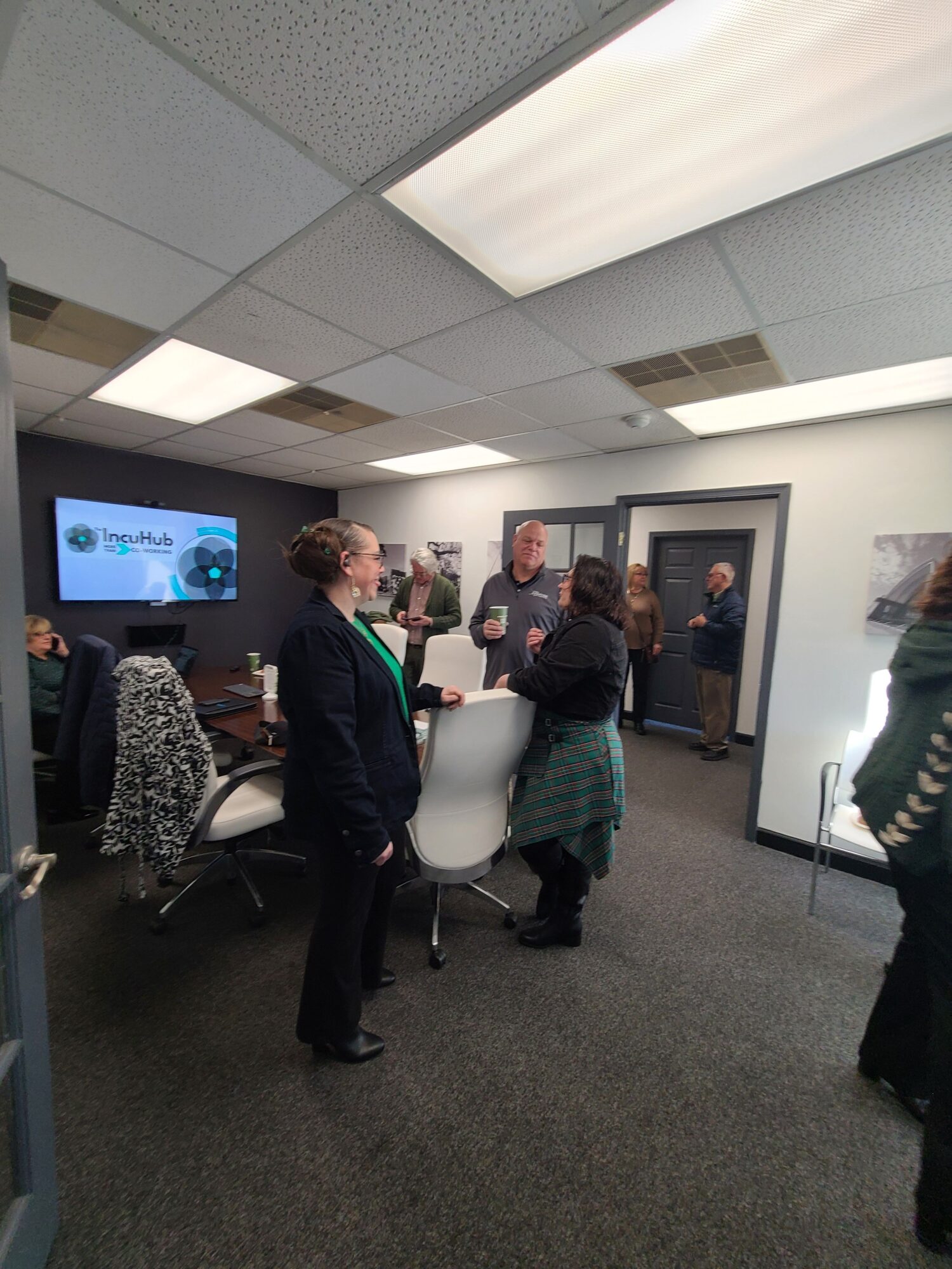 People gathered in a room with framed pictures on the wall, a large monitor, and a table with items.