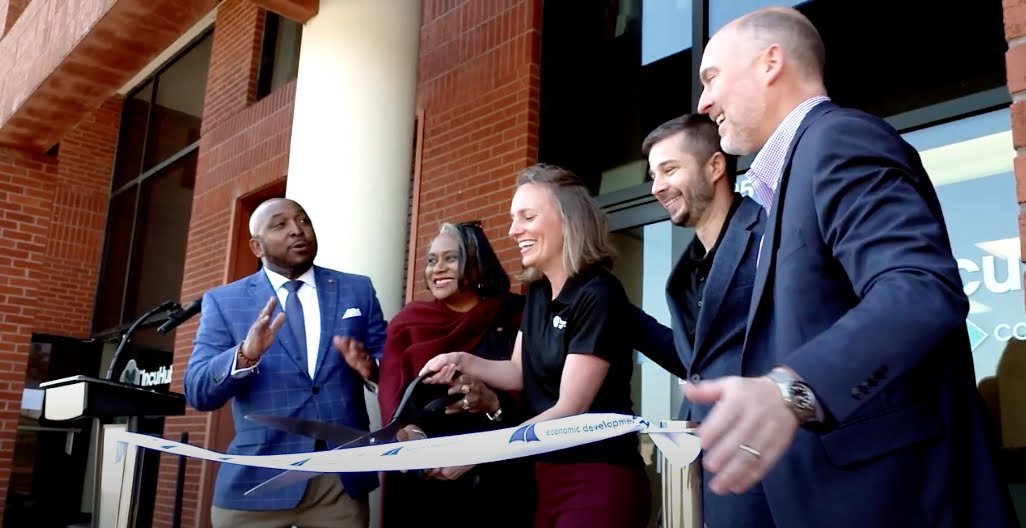 Group of five diverse people standing outside a building, smiling, and engaging in conversation during a ribbon-cutting event.