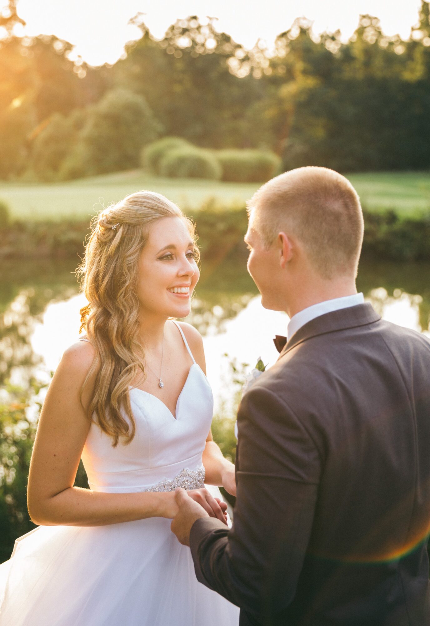 A woman in a white dress and a man in a suit hold hands outdoors, smiling at each other with a park and pond in background.