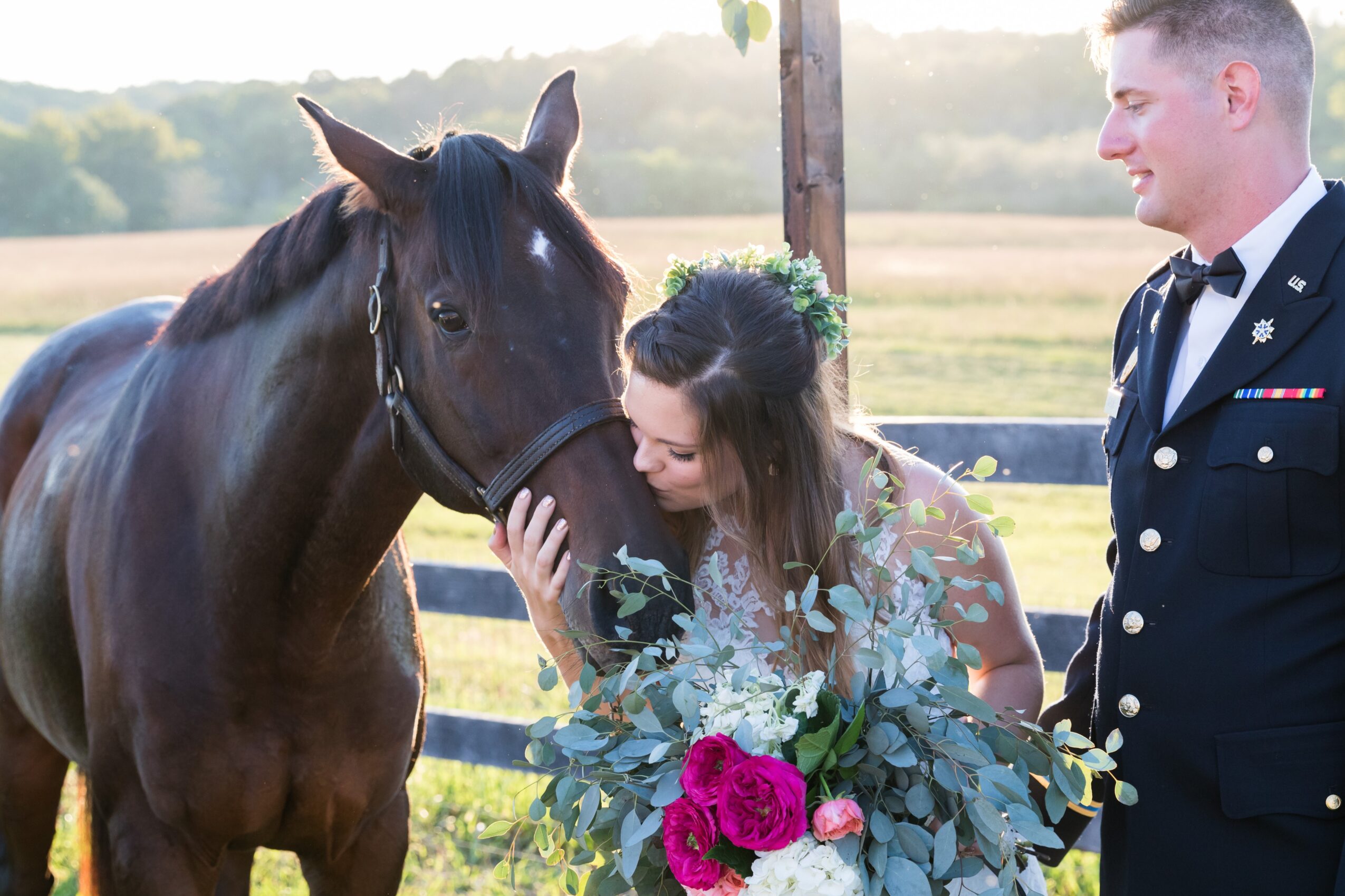 A woman in a wedding dress and a man in a military uniform stand with a horse outdoors, woman holding a bouquet of flowers.