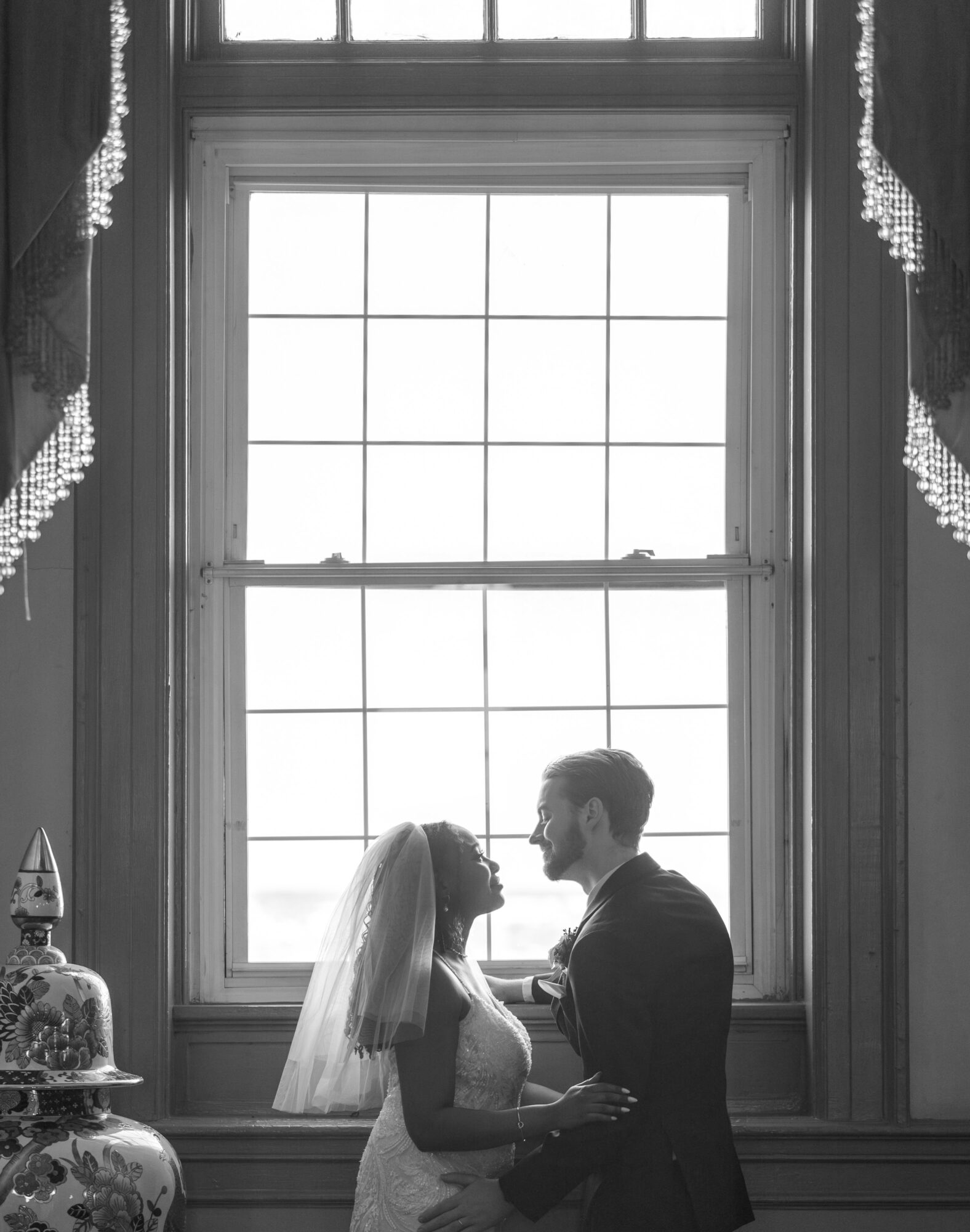 Couple standing close by a large window, holding hands and looking at each other, with curtains on sides, in black and white.