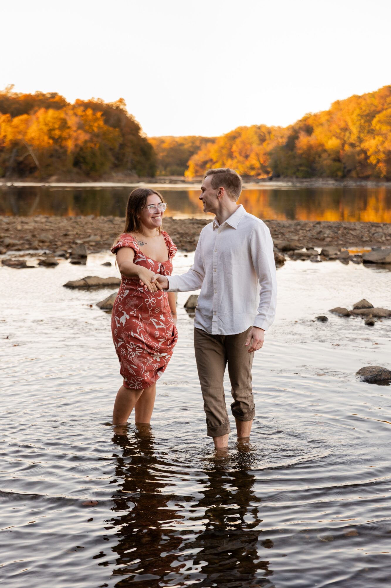 A couple stands in a river holding hands, with trees and a lake in the background during autumn sunset.