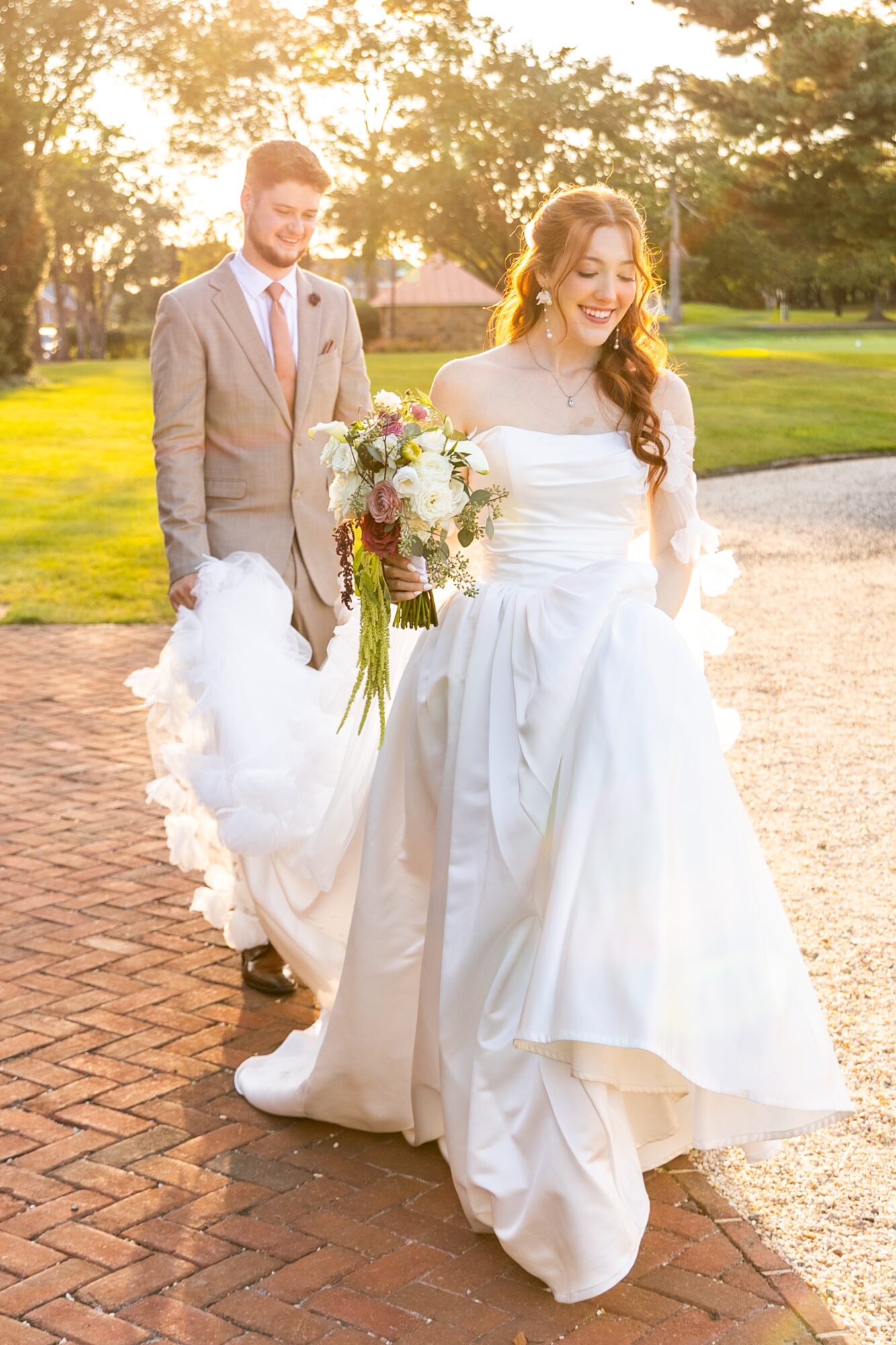 Bride in white wedding dress holding a bouquet, walking outdoors with groom in suit behind her, trees and sunlight in background.