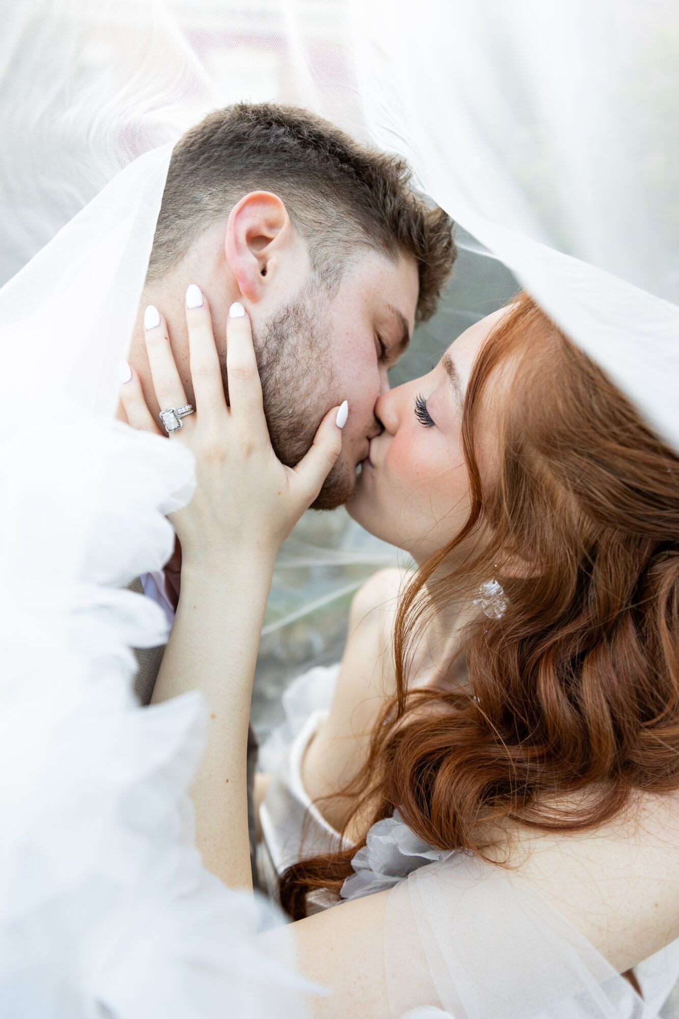 A couple with closed eyes about to kiss, holding each other's faces under a sheer fabric, in a close-up shot.