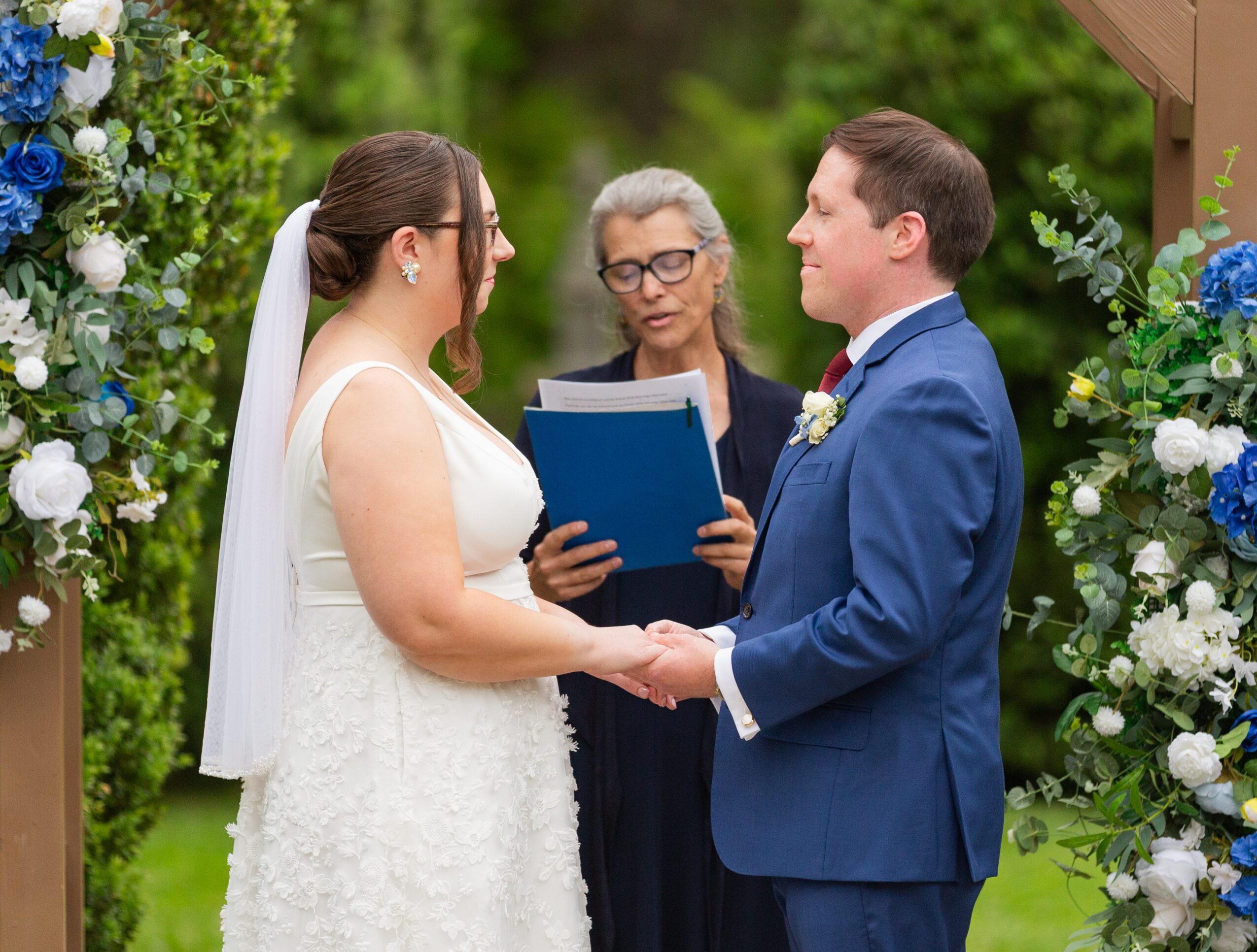 Bride and groom holding hands during wedding ceremony outdoors, officiant reading from a book, surrounded by floral decorations.