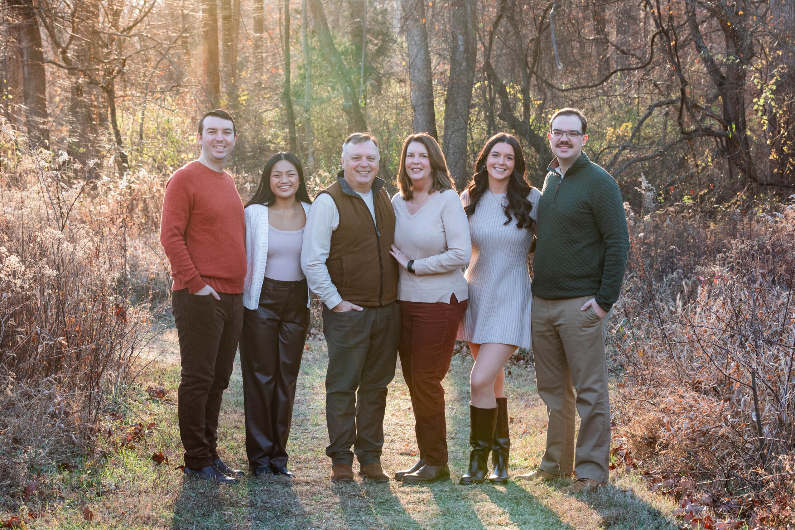 Group of seven people standing outdoors in a wooded area during autumn, smiling and posing for a photo.