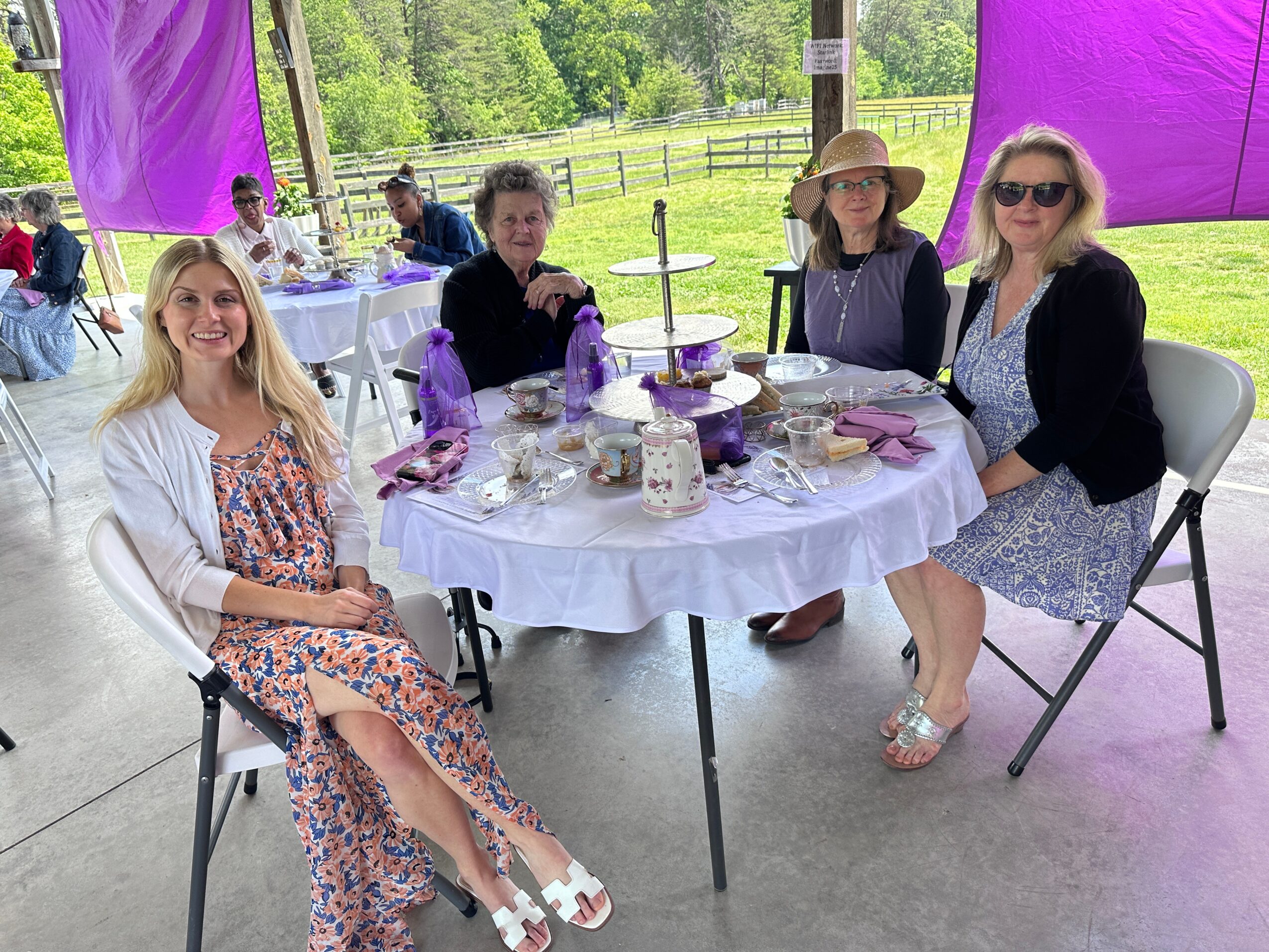 Four women sit around a table with purple decorations outdoors, smiling, with trees and a fence in the background.