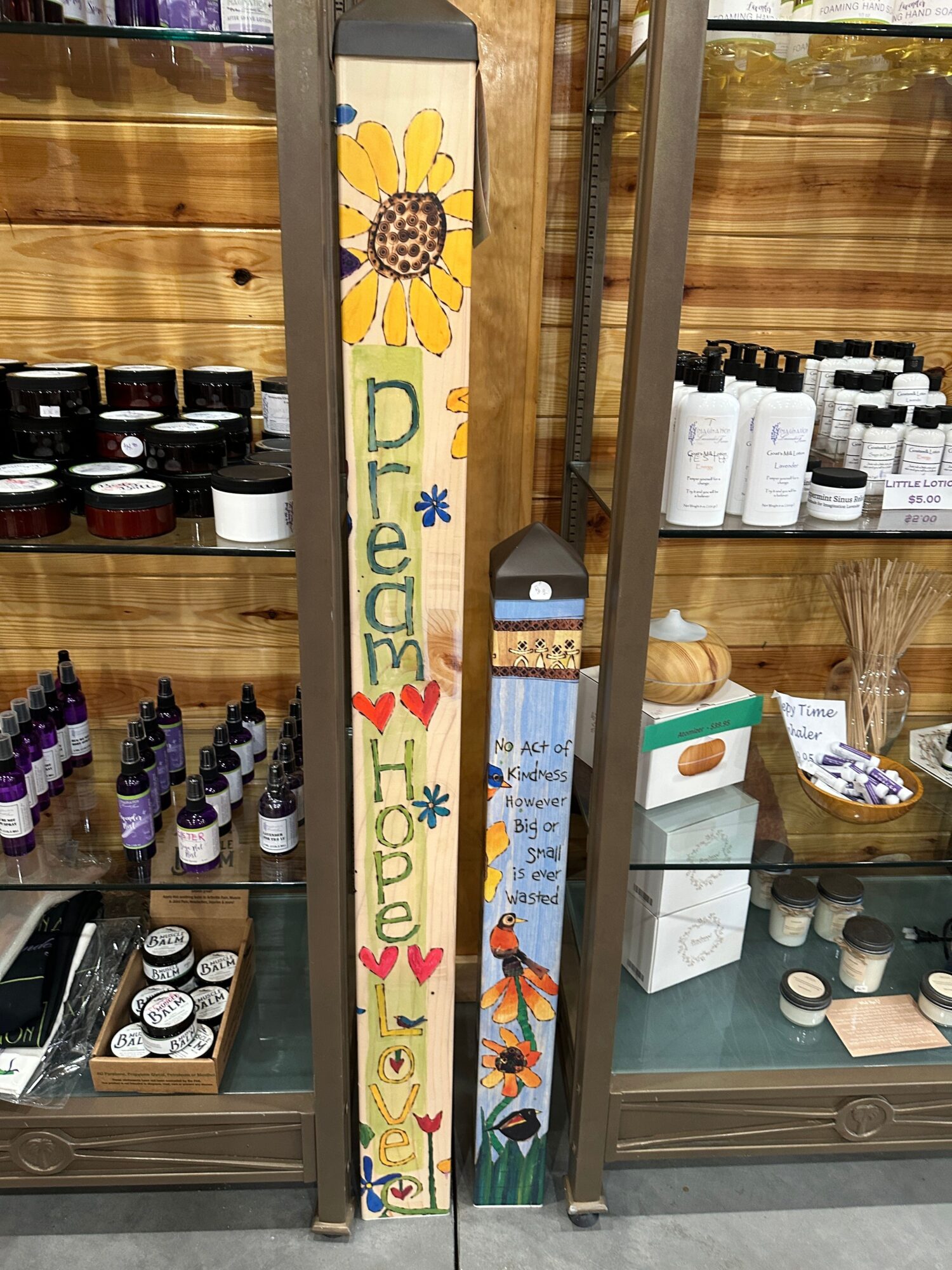 Long decorative wooden sign with colorful painted words and sunflower illustration, displayed on a shelf with various products behind glass.