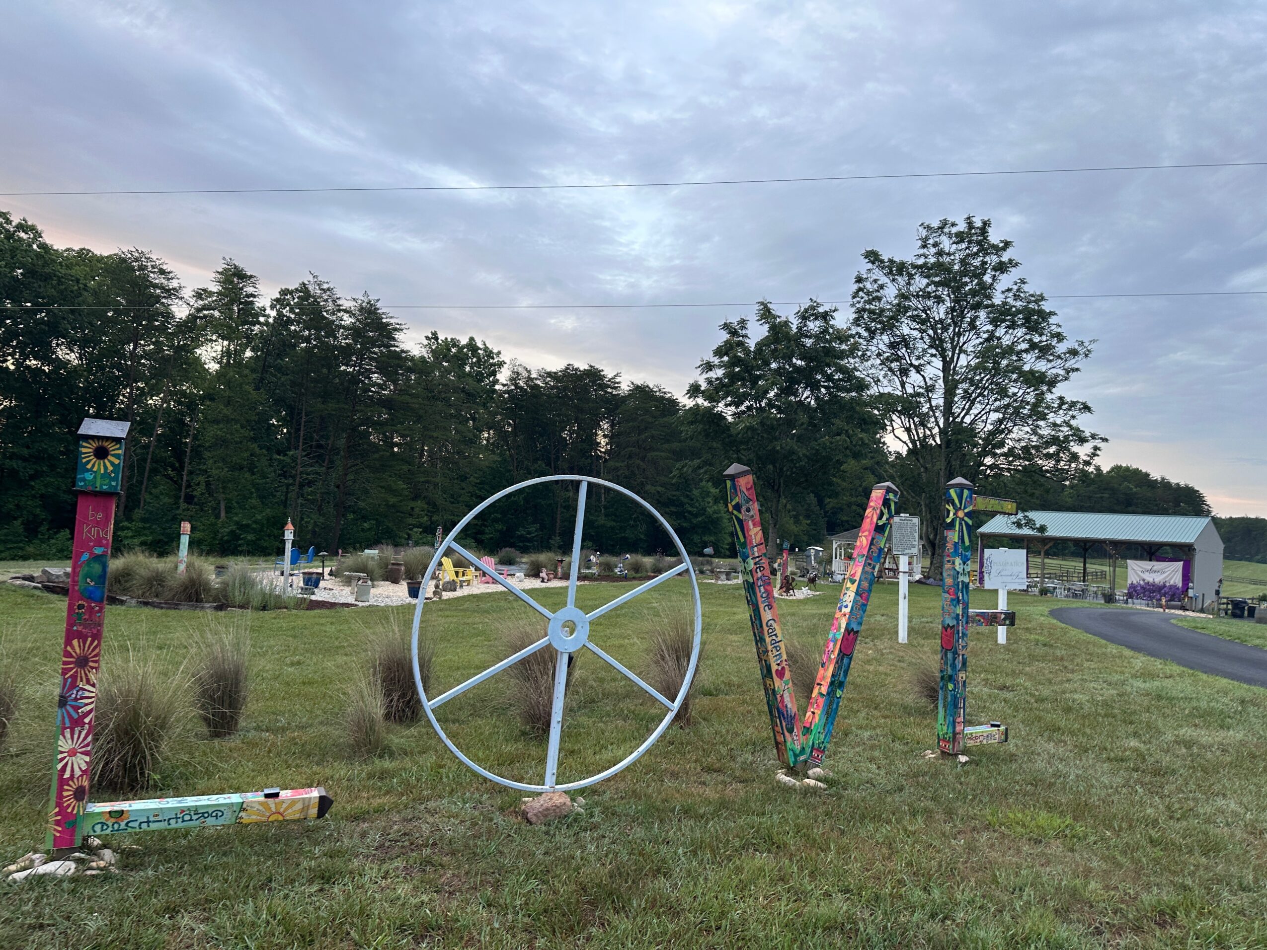 Colorful outdoor art spelling 'LOVE' with a wheel replacing the 'O' on a grassy field, trees, and a building in the background.