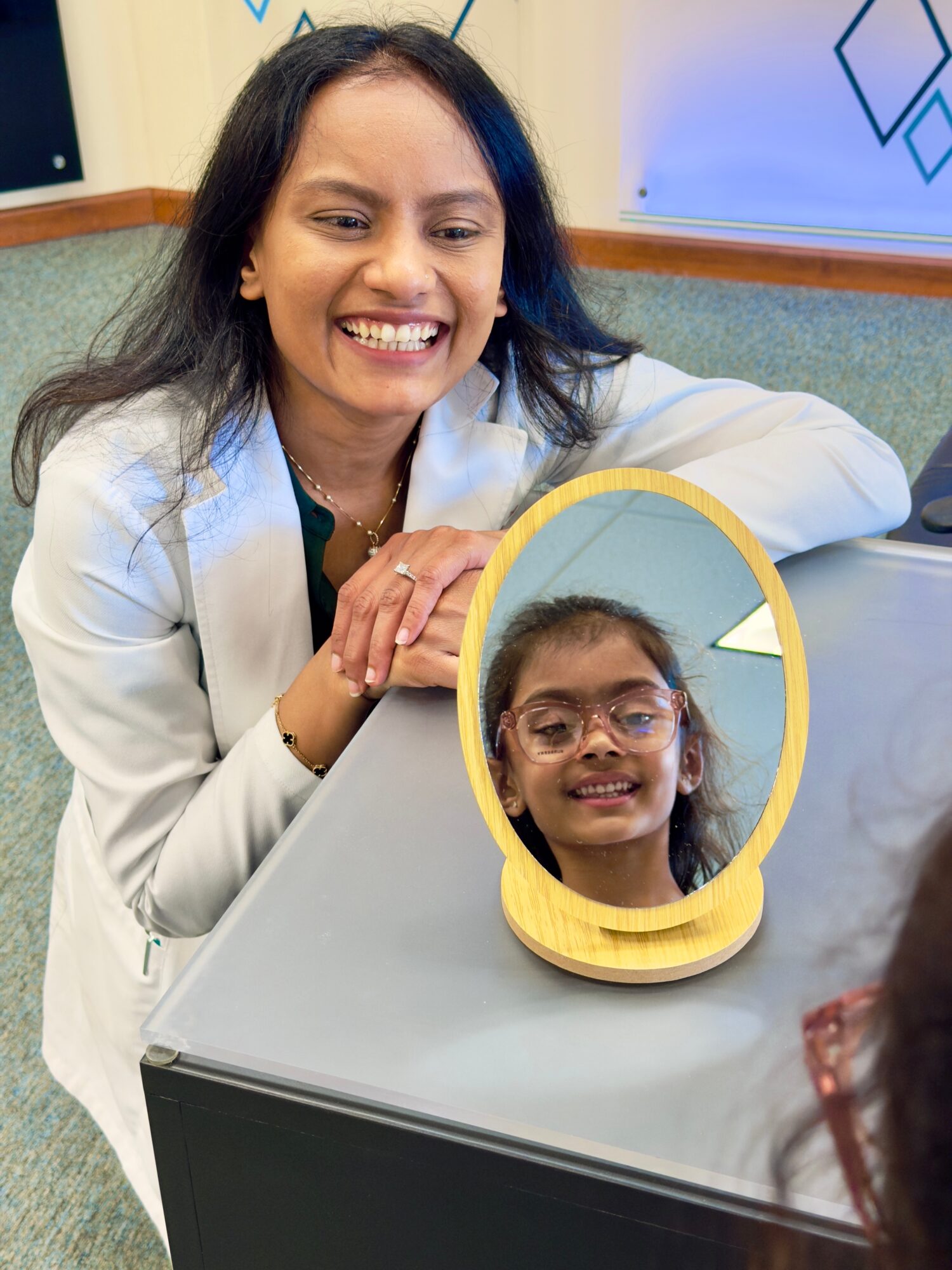 Woman smiling at mirror with a young girl’s reflection, both indoors, woman wearing white coat, girl with glasses, yellow mirror frame.