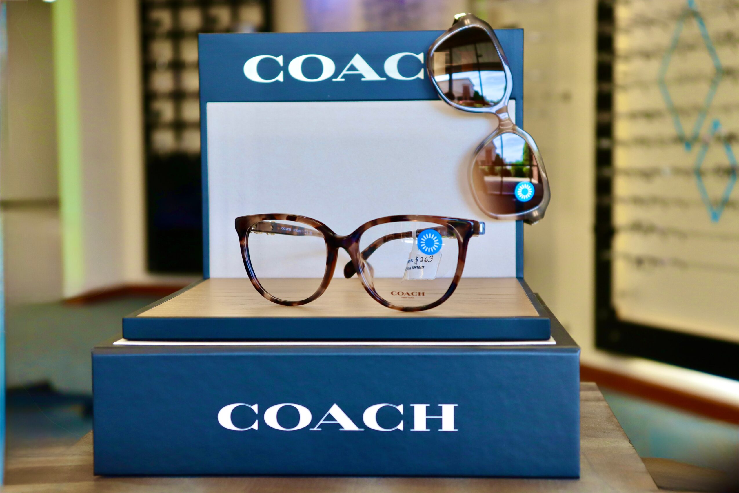Pair of sunglasses on a blue COACH display box with another pair hanging above, in a retail setting.