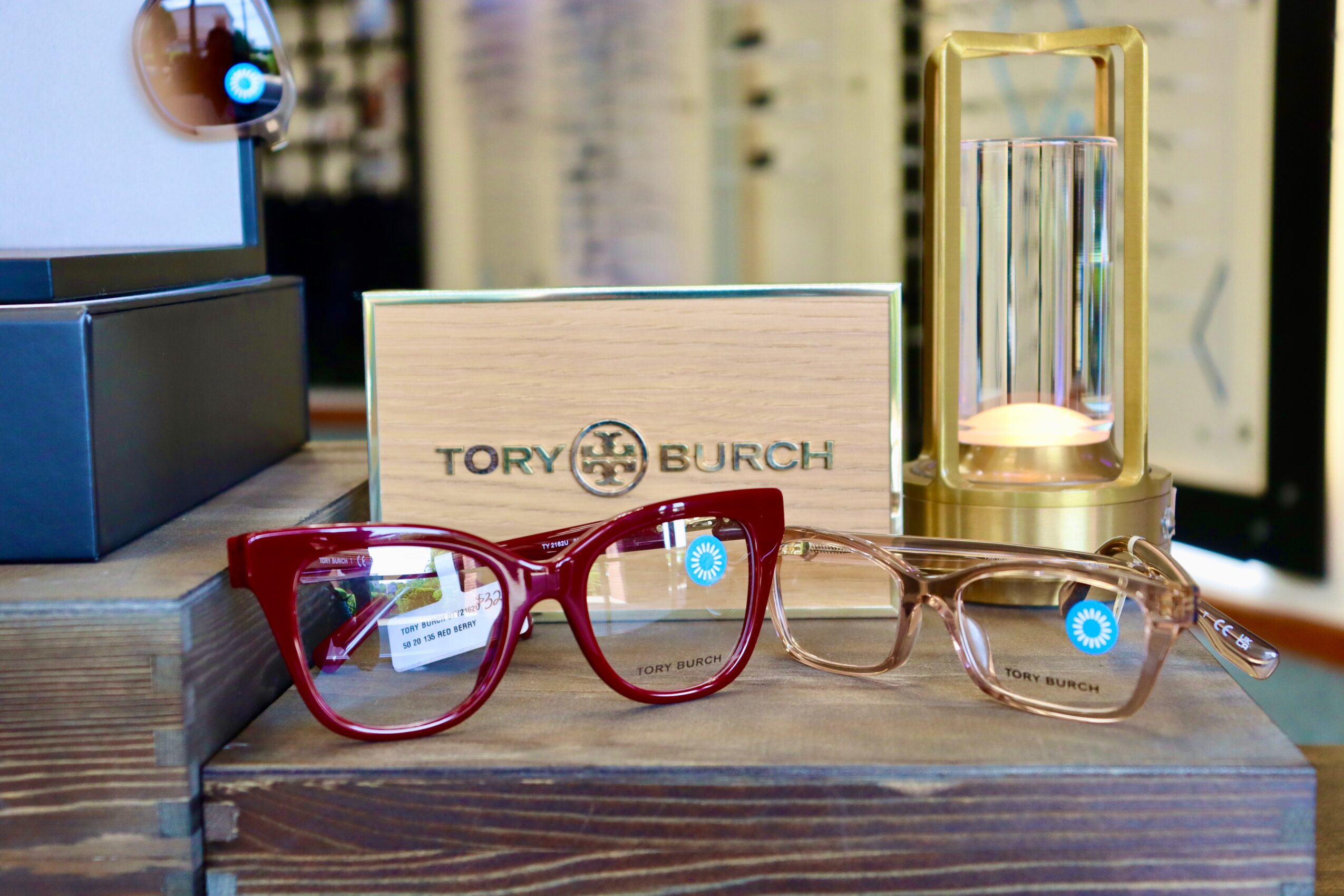 Two pairs of eyeglasses on a wooden surface with a Tory Burch sign behind them.