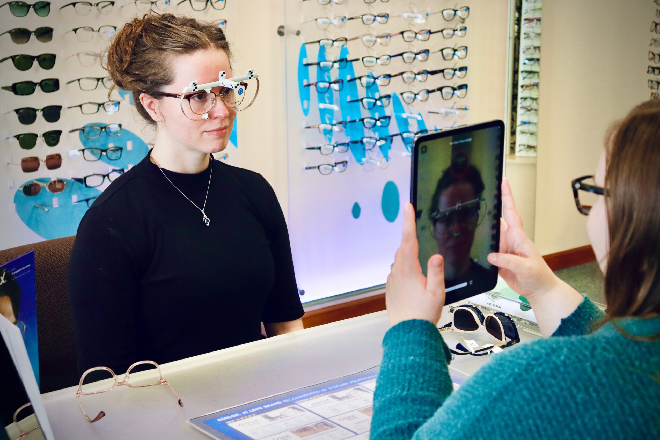 Woman with glasses and a bun poses for a photo in an eyewear store, with display of glasses behind her.