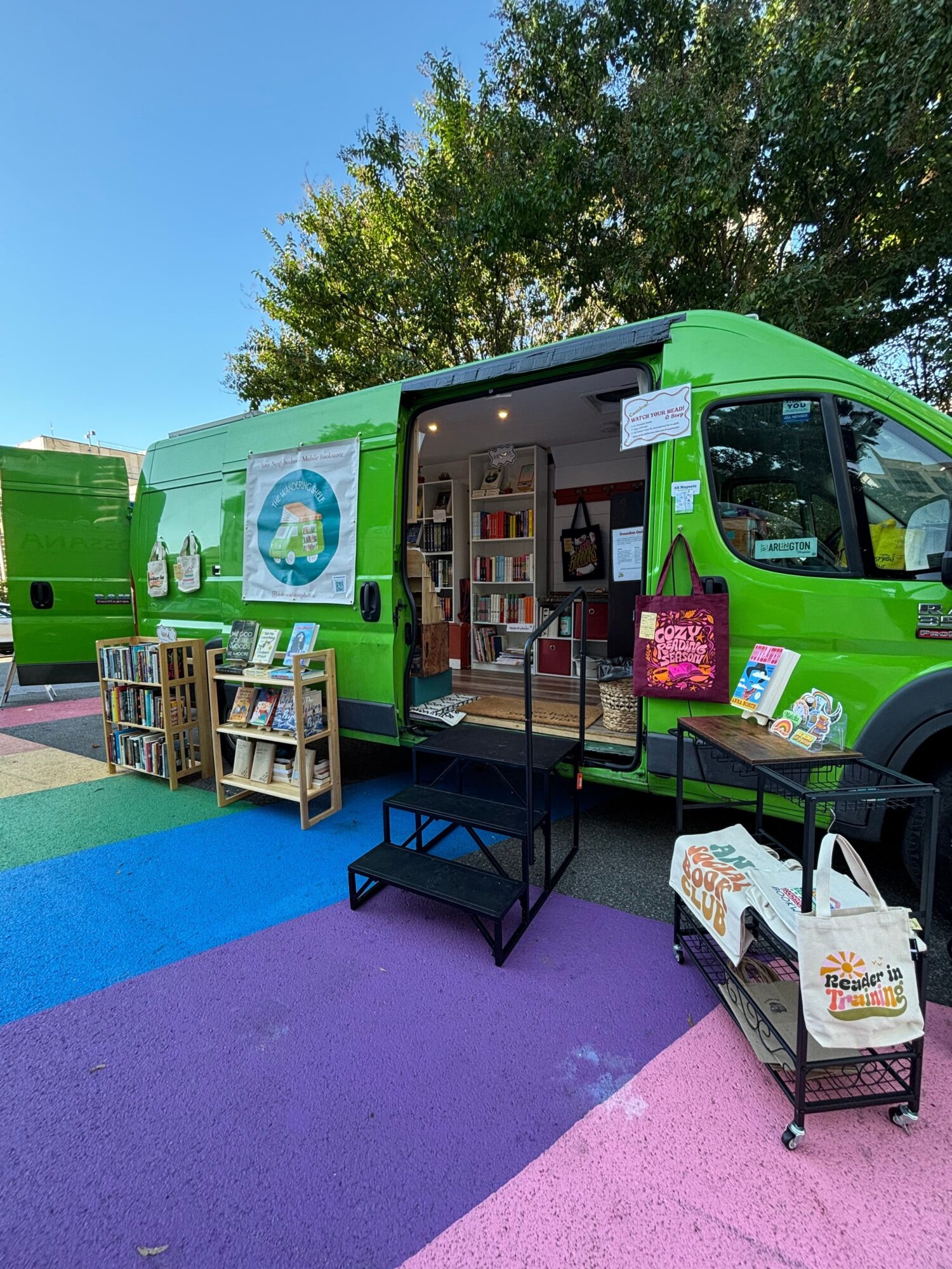 Green mobile library with books displayed outside, colorful pavement, and trees in the background.