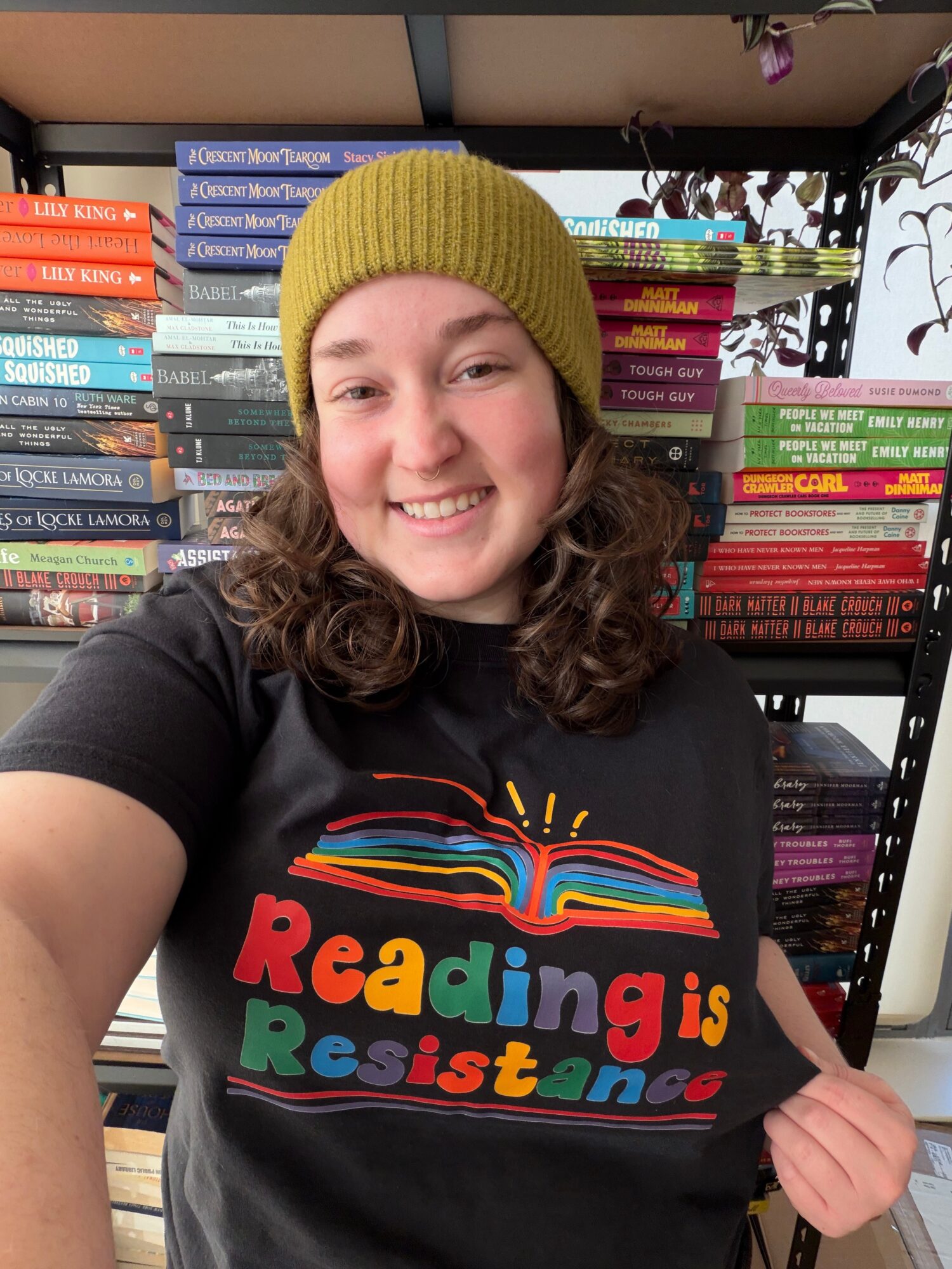 Young woman with curly hair wearing a yellow beanie, smiling, in front of bookshelves, holding a shirt with colorful text.