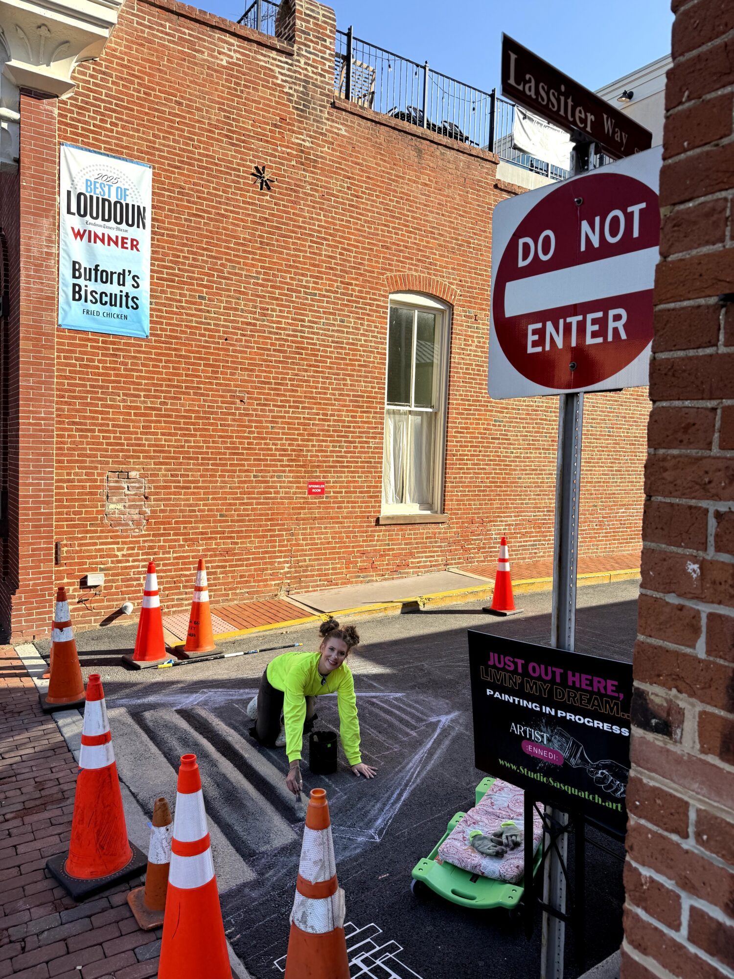 Child in neon green jacket crouching on street surrounded by orange traffic cones, with a sign and brick wall nearby.