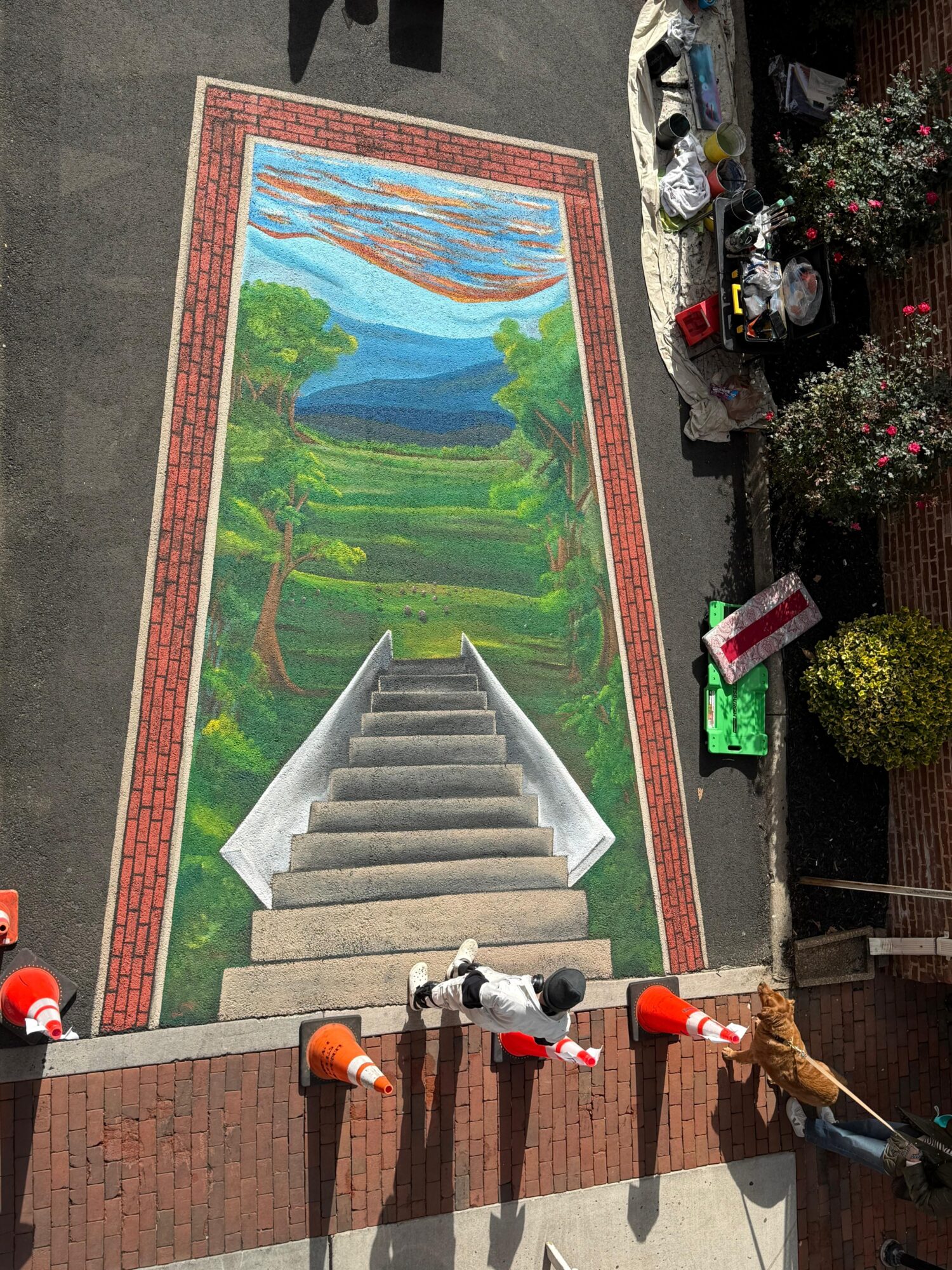 Colorful mural of a landscape with stairs leading up, trees, and a sky with clouds, painted on a street pavement.