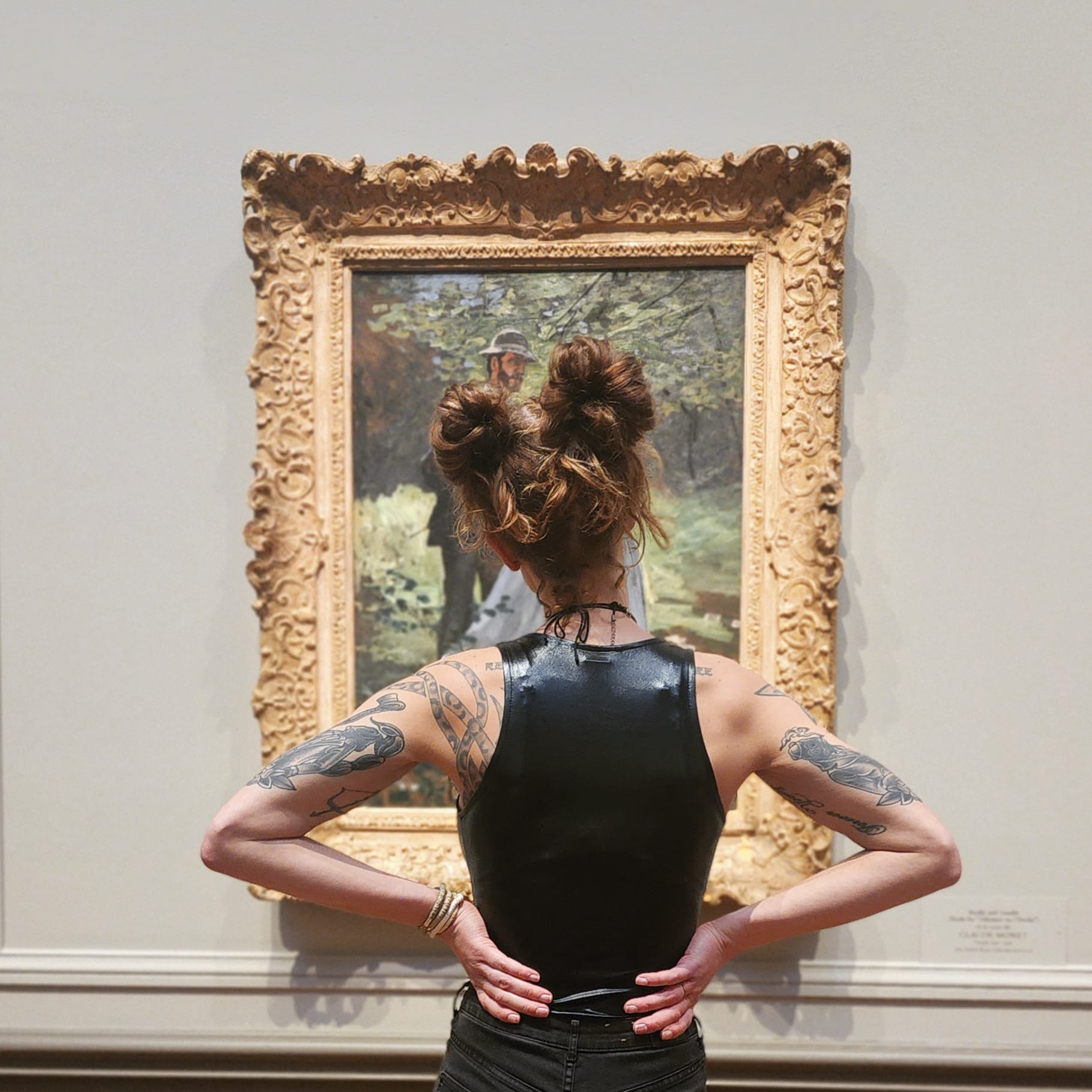 Person with tattoos and styled hair viewed from behind, standing in front of a framed painting on a wall.