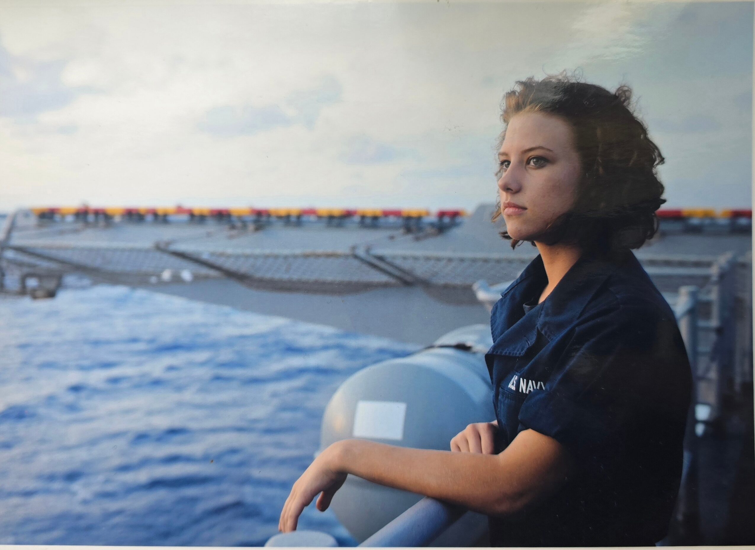 Young woman with short curly hair looking at water near a large ship, holding a railing, cloudy sky in background.