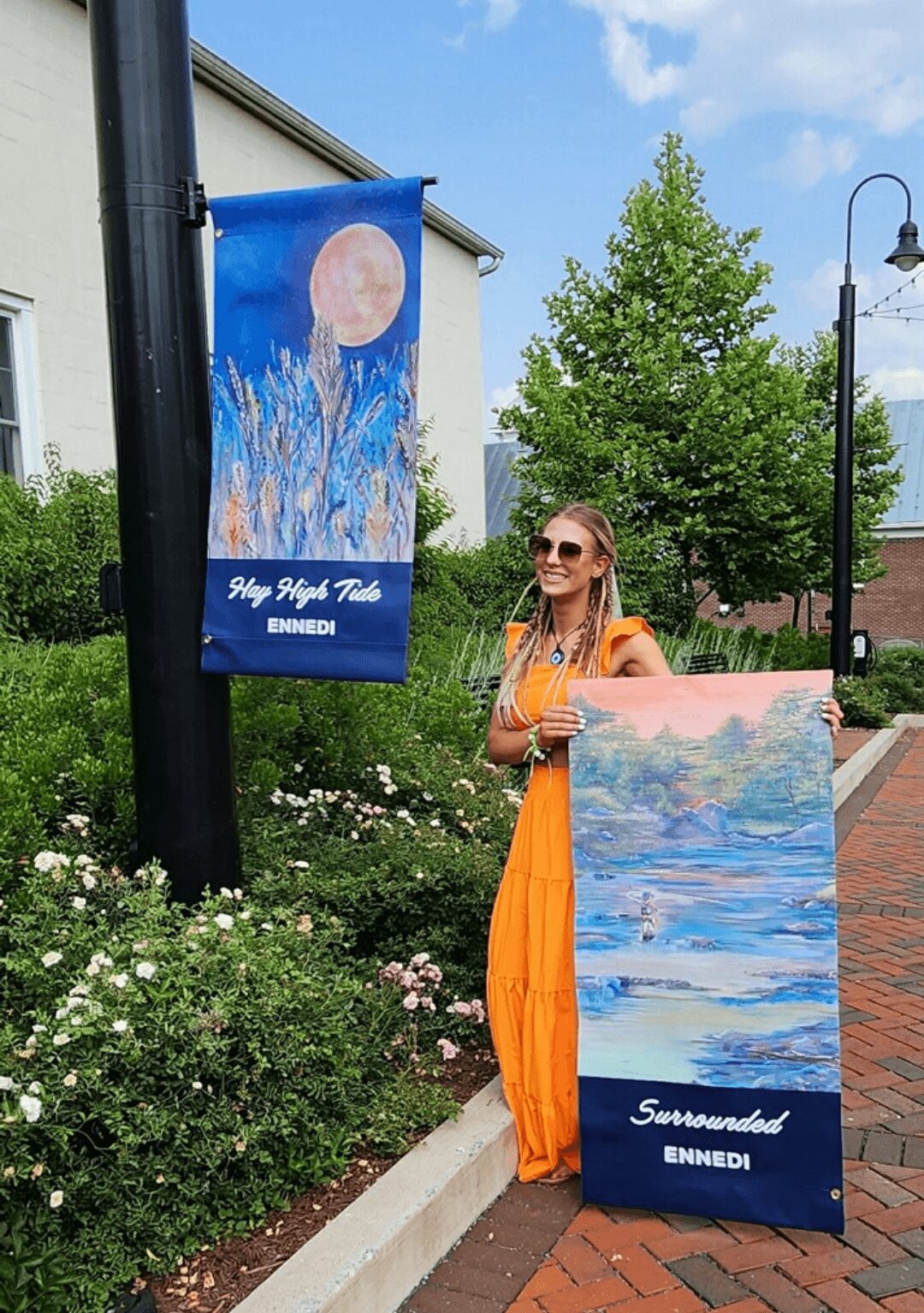 Woman in orange dress holding a large landscape painting outdoors, with banners and greenery around her.