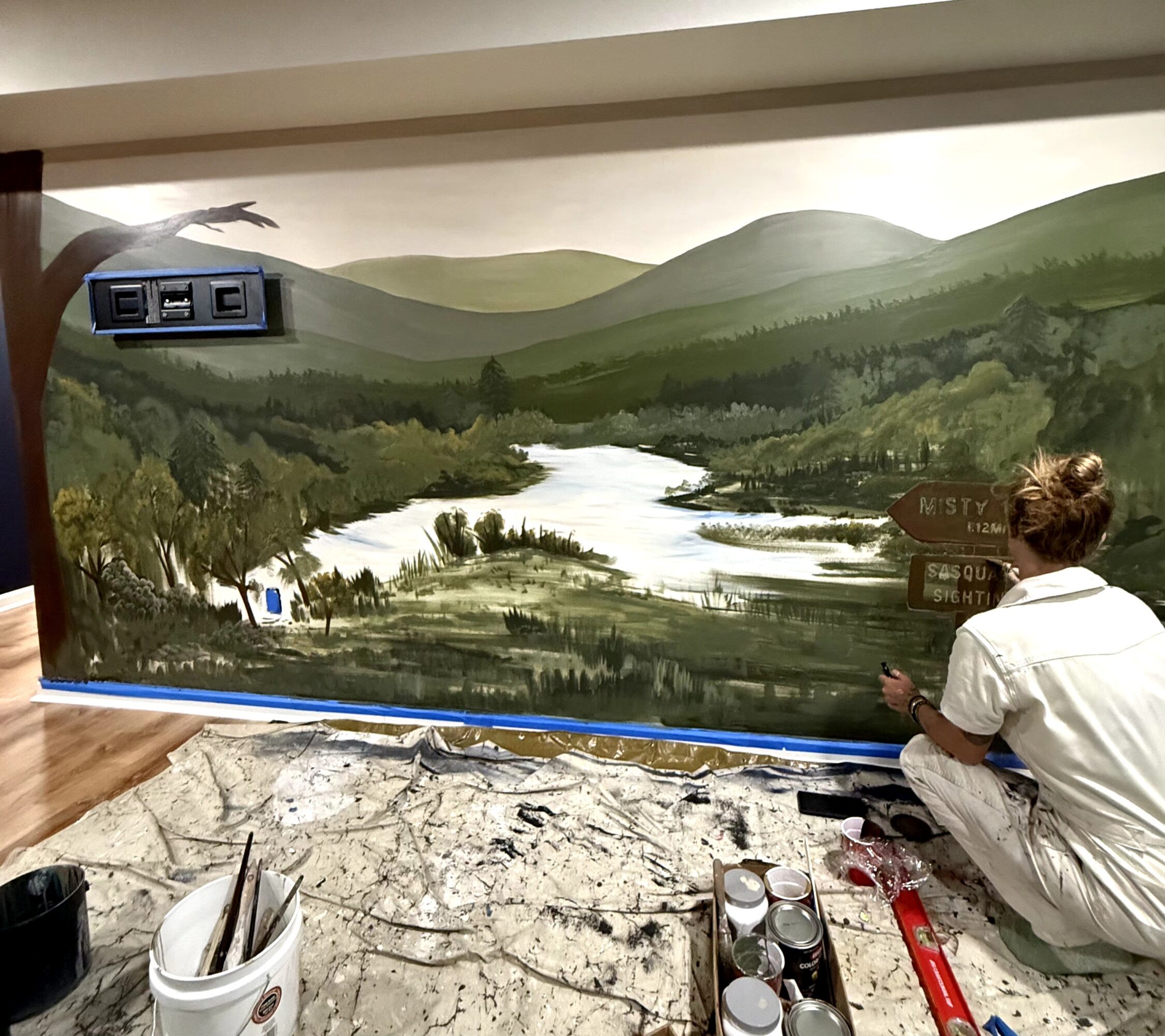 Artist painting a landscape mural of mountains, river, and trees on a wall, with painting supplies nearby.