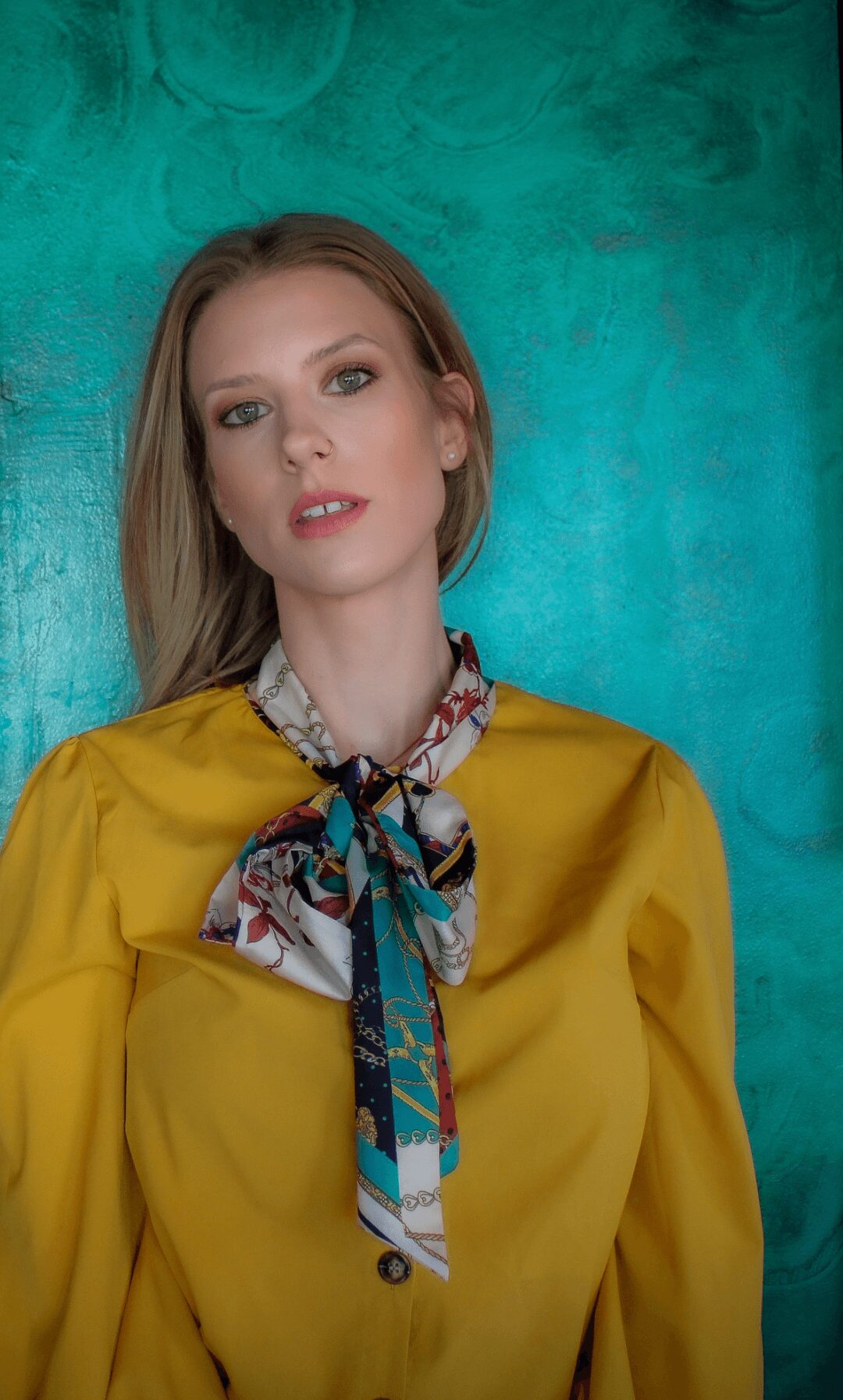 Woman with long hair wearing a yellow top and a colorful scarf against a teal background.