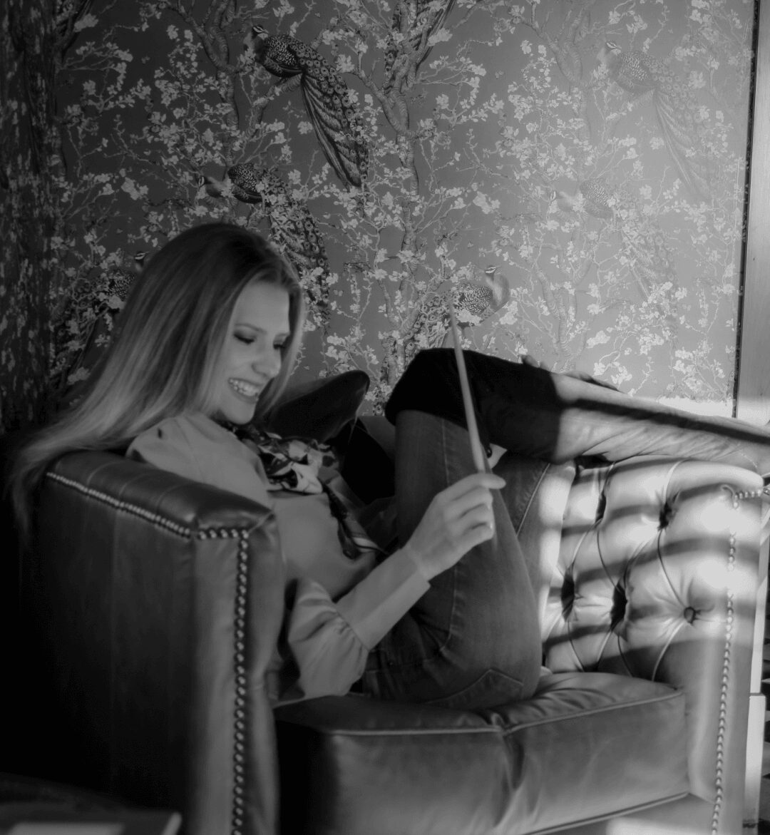 Woman sitting on a sofa, smiling, holding a stick, with a floral wallpaper background.