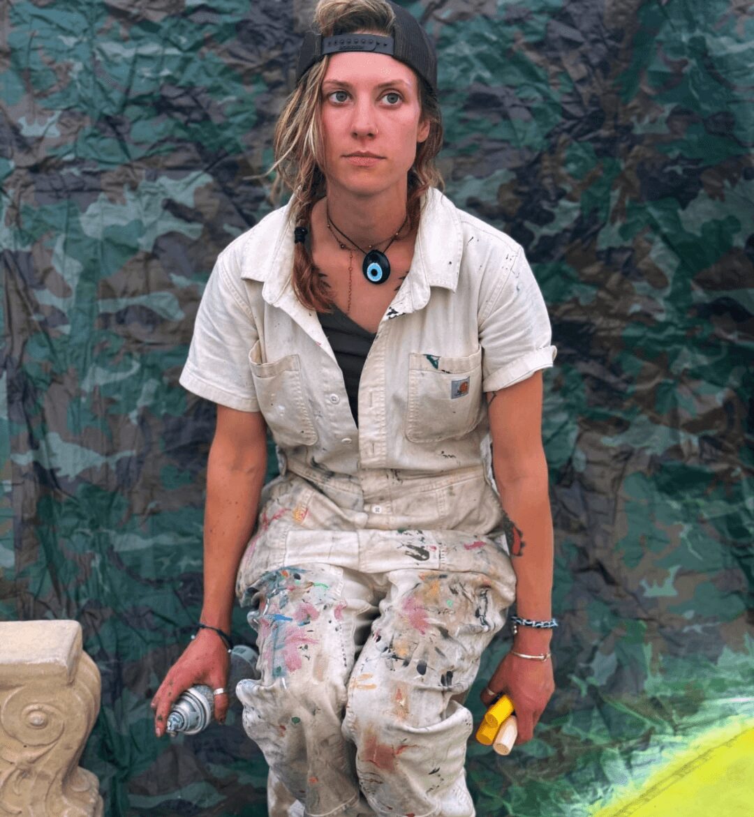 Young woman with long hair, wearing a cap, beige overalls, holding a spray can and a yellow object, standing in front of a camouflage background.